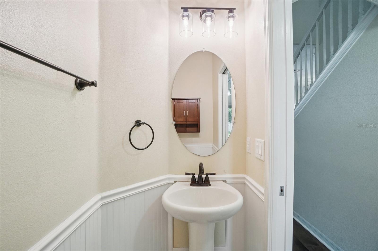 Half Bath - The downstairs half bath features a classic pedestal sink with rubbed bronze faucet and fixtures, an oval mirror, modern vanity light bar, and white beadboard wainscoting — a clean, well-appointed powder room for guests.