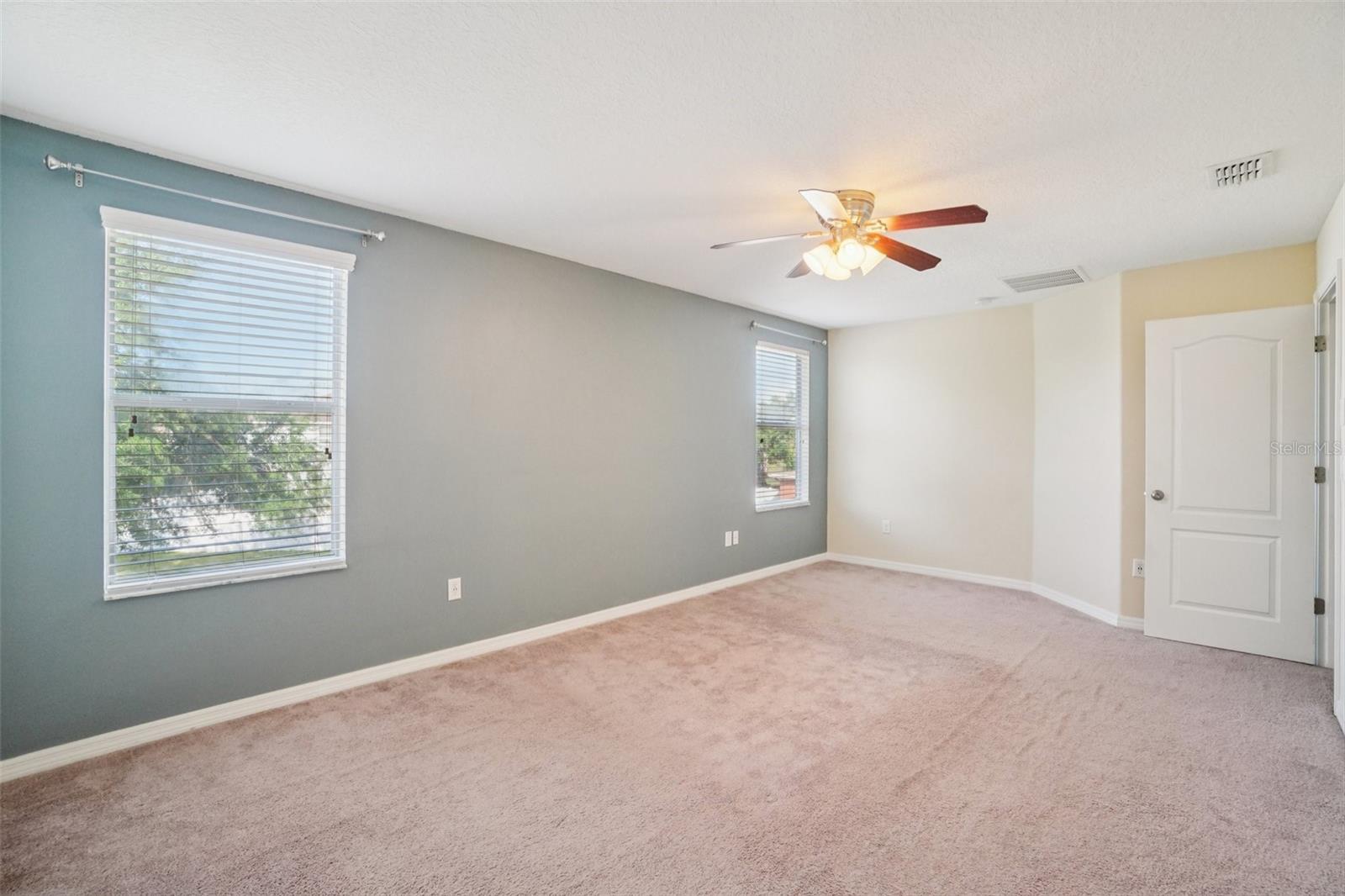 Primary BedroomThe spacious primary bedroom features plush carpet, a ceiling fan, two windows with natural light, and a slate blue accent wall — a bright, well-appointed retreat with ample room to unwind.