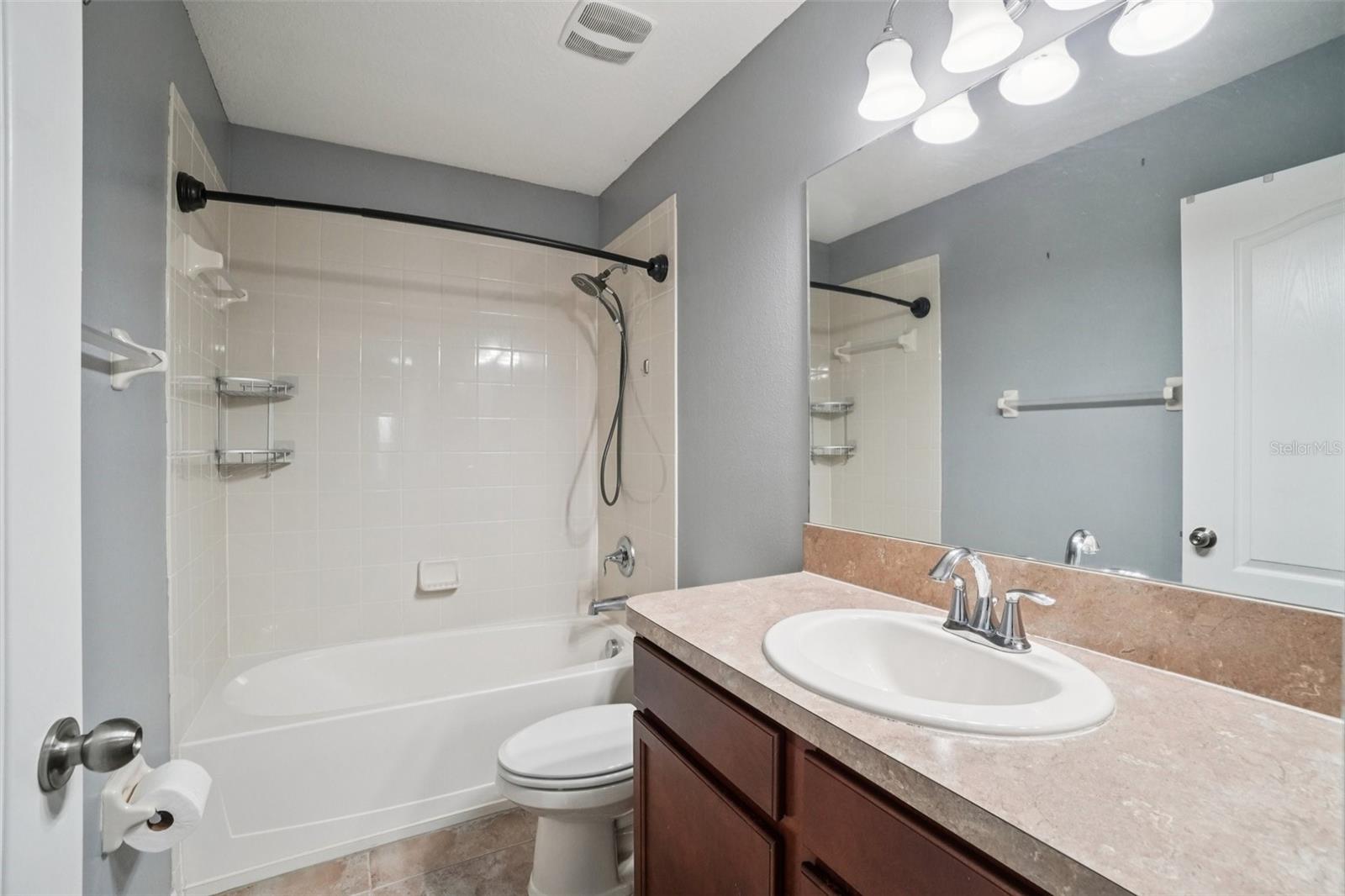 Hall BathroomThe upstairs hall bathroom features a single vanity with dark cherry cabinetry, tile countertop, large mirror, multi-bulb light fixture, tub/shower combination with tile surround, and large-format tile flooring — clean and well-appointed for everyday use.