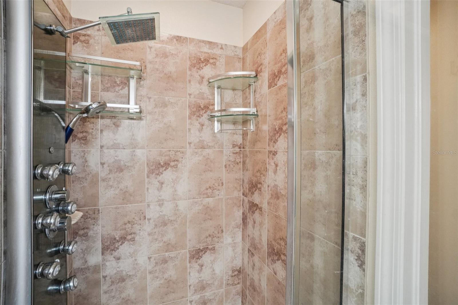 Primary shower with travertine-style tile surround, rain showerhead, multiple body jets, dual glass corner shelves, and frameless glass enclosure.