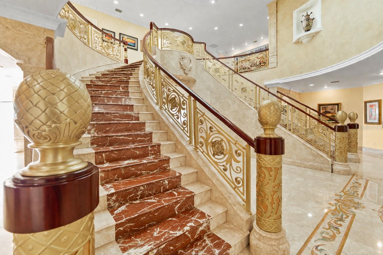 Sweeping dual marble inlaid staircases with gold and iron balustrades and ornate railings.
