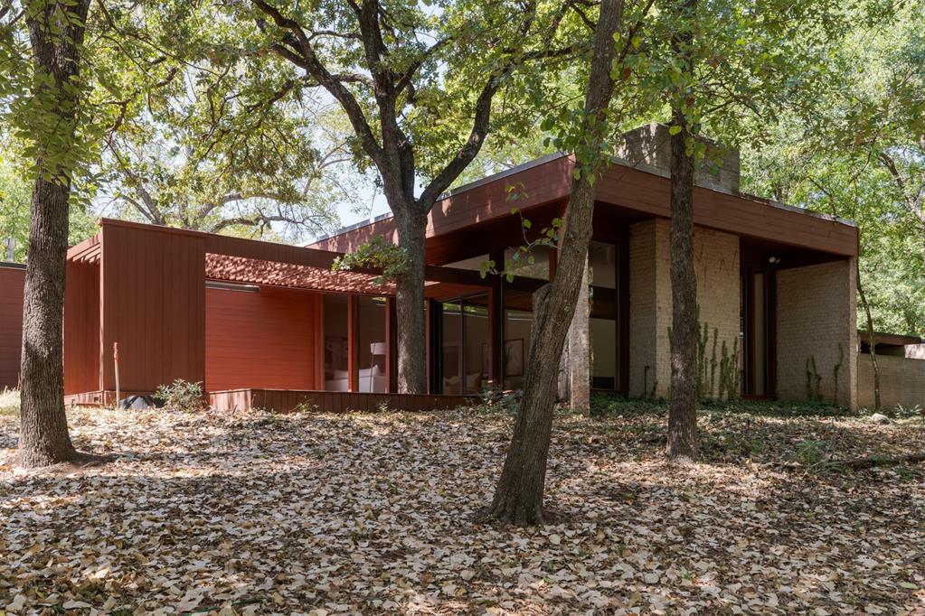 Designed with Northern California architectural influence and brought to life in 1980, this single-story home sits on 5.94 private, densely wooded acres — a scale and seclusion that simply don't exist elsewhere in this price range in the Edmond market. A long private drive winds through mature oak canopy to a home that reads more Sea Ranch than suburban Oklahoma: deep roof overhangs, board-and-batten siding, and repeating slatted wood pergolas carried from exterior to interior. Walls of glass on nearly every elevation erase the line between the house and the forest around it, with clerestory windows throughout the west that flood the home with light while preserving privacy. This is Organic Modernism, genuinely transplanted, on acreage most buyers assume doesn't exist this close to the city. A true architect-designed residence with post-and-beam construction, terrazzo flooring in the main living areas, and hand-detailed wood ceiling treatments that give it a one-of-one presence. The great room centers on a whitewashed brick fireplace wall beneath an exposed timber beam, and a dedicated library wall of built-in shelving speaks to serious original craftsmanship. The kitchen features a full stainless appliance package and dual granite islands, opening directly to a light-filled dining area.The primary suite stands out with dual his-and-her baths — one finished in a mother-of-pearl tile mirror surround with blown-glass pendant lighting, the other a bold cobalt-tiled shower — plus a jetted soaking tub set beneath a private skylight with direct deck access. A separate in-law suite with its own kitchenette, living room, and private entrance offers flexibility for multi-generational living or extended guests. Nearly six acres of mature trees and genuine seclusion minutes from I-35, Broadway Extension and the Turner Turnpike. This is a home for a buyer seeking architectural significance, real privacy, and acreage close to the city that is virtually impossible to replicate.