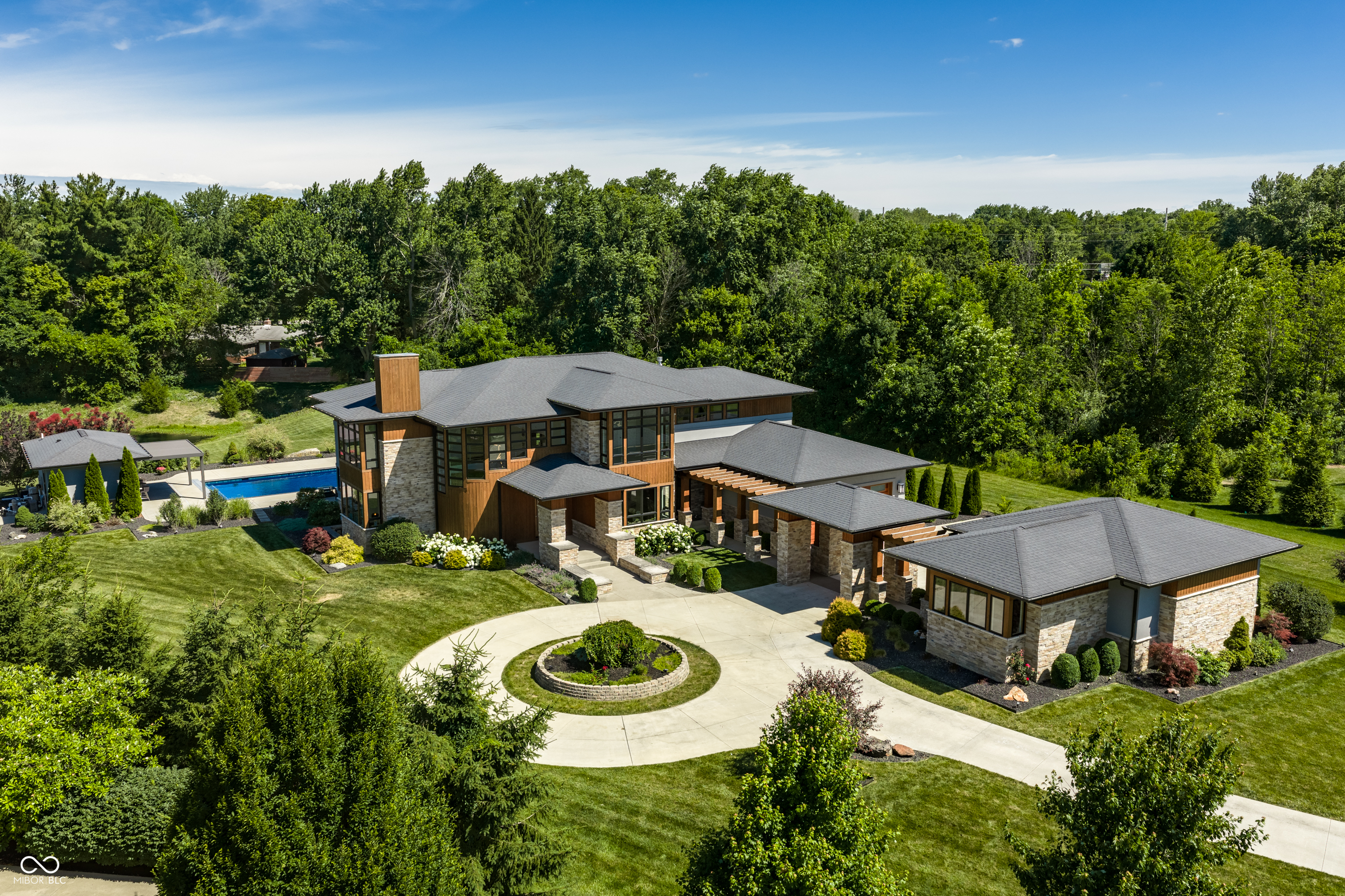 6301 Montana Springs Drive is a G&G Custom Homes masterpiece set back on 2.44 private acres in Shannon Springs, where organic luxury meets warm cedar, stacked stone, and black framed windows beneath dramatic rooflines that create an exterior presence that is unmistakably its own. A long sweeping driveway ends at a timber pergola walkway that connects the home's structures, an arrival that sets the tone for everything that follows. Inside, large stone pillars and a striking fireplace define the great room, where two-story windows blur the line between indoors and the private grounds beyond. A dramatic upper catwalk looks down over the space, adding a sense of scale that is immediately felt. An impressive chef's kitchen with custom walnut cabinetry opens to the great room and breakfast nook for seamless entertaining. A walk-through pantry with open shelving connects the kitchen to the mudroom on one side and a formal dining room on the other, offering ample space for gathering at every hour of the day. The main level also offers a bedroom suite with its own porch, a dedicated office, and a curved staircase with skylight rising to the upper level. Three bedrooms upstairs include a primary suite with a spa-like bath, California Closets, and a private deck, alongside a shared loft that invites relaxation. The lower level offers a bar, theater, private bedroom, and lounge, along with a generous unfinished space with egress windows offering room to grow. A fully redone gunite pool and hot tub reveal a backyard that feels like a resort of its own. A pergola covers fireside seating around a double-sided fireplace and built-in island, grass pavers extend into additional gathering space, and a full-service pool house with bar and bathroom completes the setting, all overlooking a tranquil pond and wooded treeline. Private, intentional, and unlike anything else in Zionsville, this is a property that feels worlds away while remaining close to everything the town has to offer.