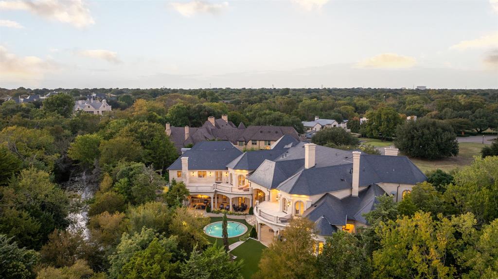 Welcome to 10617 Bridge Hollow — a masterwork of architecture, integrity + serenity. Tucked in the exclusive Creeks of Preston Hollow, this residence rests on a private lot where 2 creeks wind through lush, tree-lined banks. It stands as a rare union of natural beauty + enduring craftsmanship. This 2018 custom home spans 1.44 AC + is framed entirely in steel — a $500K investment ensuring lifelong stability. 130+ piers run deep into Austin chalk, forming a foundation that’s a feat of engineering. The timeless design features a Syrian limestone façade + slate roof built for a century of endurance. Hand-carved fireplaces, steel gutters + integrated drainage reveal an obsession w permanence + precision. Step through the 20-ft African mahogany door + the home unfolds in a play of light + volume. Ceilings soar to 25 ft in the entry + 17 in the living room, framed by windows overlooking nature. Each space exudes quiet grandeur — oak + limestone floors, bespoke chandeliers + refined proportions. The kitchen is a sculptural statement in quartzite, onyx + brass, backed by a scullery for effortless entertaining. The Control4 system unites lighting, sound + security. The 1st-flr owner’s suite is a private sanctuary w heated marble floors, automated shades, massive closets + a massage-exercise room open to a serene backyard. A library w built-ins + a custom fish tank adds sophistication + warmth. A spacious guest suite rounds out the main level. Upstairs, 4 guest suites w private baths overlook treetops + water views, while a glass staircase from Australia arcs through the home like sculpture. A game room offers flexible space for entertaining + a basement wine cellar provides abundant storage for collectors. A resort-style backyard, inspired by Palm Springs, features a tranquil pool, turfed lawn + elegant lighting — a landscape of privacy + peace. This residence is a testament to enduring design — built for those who value substance, beauty + permanence in equal measure.