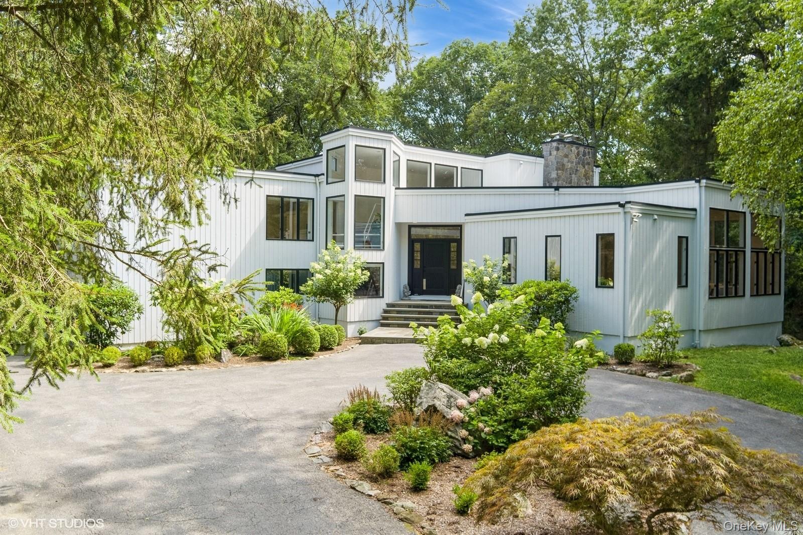 Welcome to this sleek, light-filled Contemporary in Armonk (North Castle), where openness and design converge on 2.21 private acres in the award-winning Byram Hills school district. A circular driveway leads to a dramatic double-height foyer with gleaming marble floors, setting the tone for the sun-drenched interiors that follow.
Walls of windows, soaring ceilings, and skylights flood the home with natural light. The expansive formal living room features a striking floor-to-ceiling stone fireplace and flows effortlessly into the spacious family room, dining area, and designer Leicht kitchen. A convenient home office is tucked just off the kitchen. Sliding doors open to a wraparound deck that seamlessly extends the living space outdoors, perfect for entertaining or relaxing in privacy.
The private primary suite is a true retreat, complete with a balcony, two oversized walk-in closets, a dedicated shoe closet, and a generous dressing area. The spa-like bath offers a soaking tub and a freestanding shower.
Upstairs, you’ll find an en-suite bedroom plus two additional bedrooms that share a hall bath. The walkout lower level offers a vast, welcoming space with designated areas for recreation, fitness, media, and guests, plus a full bath, cedar closet, and abundant storage.
With multiple fireplaces, flowing open spaces, and serene outdoor surroundings, this home offers a harmonious blend of contemporary design and everyday comfort.