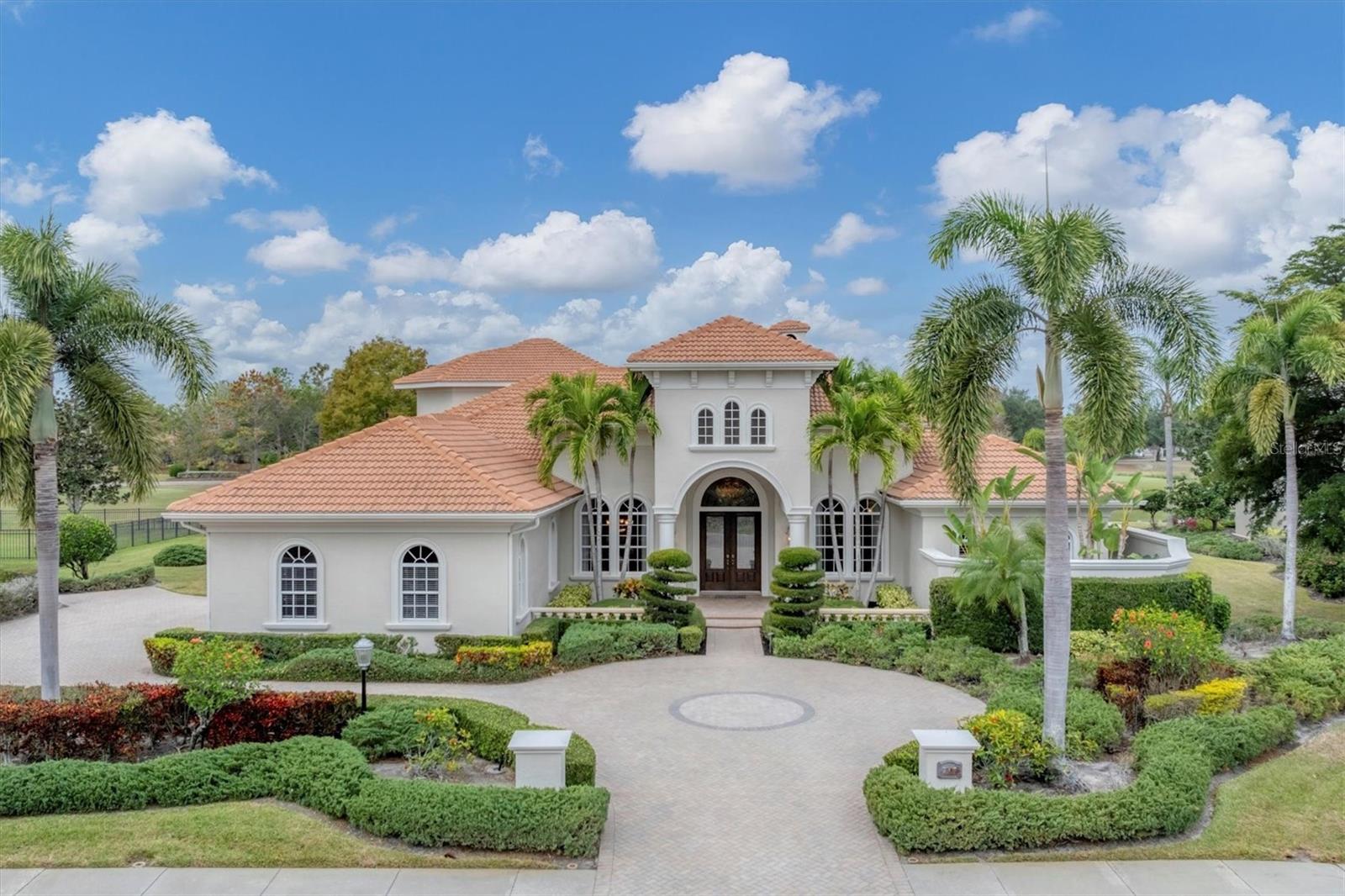 Welcome to the Anchor Builders Grand Cypress Model, a timeless estate residence set in the prestigious Lakewood Ranch Country Club, ideally on desirable Portmarnock Place. Resting on a rare half-acre waterfront homesite with sweeping views of the Cypress Links Golf Course, this home embodies refined Florida living at its finest. A grand portico entrance, decorative paver driveway and impeccably manicured tropical landscaping create a commanding first impression. Inside, soaring ceilings and exquisite architectural detailing abound, including double crown moldings, graceful arches, intricate Corinthian columns, wainscoting, coffered and tray ceilings with uplighting, marble floors, plantation shutters and designer lighting throughout. Double wooden doors with detailed glass inlay open to a formal dining room, intimate sitting room ideal for a piano, complete with built-in shelving, and elegant living room with a wet bar with beverage cooler and a stone gas fireplace with an elegant mantle. Designed for grand entertaining and everyday comfort, the home offers four bedrooms, each with a designated bath, plus a spacious den featuring custom wood built-ins, bookshelves, and a wet bar overlooking the pool and lanai. The gourmet kitchen is a chef’s dream, showcasing abundant maple cabinetry with pullouts, granite countertops, a large island with prep sink, Viking six-burner range with double oven, custom stone vent hood, pot filler, hidden walk-in pantry, Sub-Zero and Bosch appliances, and under-cabinet lighting. An eat-in dining area with a built-in banquette enjoys stunning lanai views through oversized aquarium windows. The expansive great room flows seamlessly from the kitchen and features disappearing impact sliders, built-in media center, surround sound and smart home audio. Upstairs, a private bedroom suite or multipurpose retreat offers wood-inlaid ceilings, an elegant wet bar, balcony and en-suite bath, ideal for guests or a refined lounge. An iron-gated under stair wine cellar adds a distinctive touch of sophistication. The primary suite is a luxurious sanctuary overlooking the pool, with a spa-inspired bath featuring a garden Jacuzzi tub, walk-in shower, unique dual private areas and massive walk-in closets with a dedicated dressing area. Two first-floor guest suites showcase decorative tray ceilings, custom trim work, window seats and built-in closet storage. Outdoors, the resort-style paver lanai is exceptional, offering a gas fireplace, wood-inlaid ceiling, outdoor kitchen, saltwater pool and spa with smart control, ideal for year-round entertaining in Florida style. Additional highlights include Sonos surround sound in the kitchen, great room, living room and lanai, a side-turned three-car garage with extensive built-in storage, and thoughtful updates including a new roof (2018), all HVAC replaced, rescreened lanai (2022) and new water heater (2022). This extraordinary residence seamlessly blends architectural grandeur, modern comfort and an unrivaled country club lifestyle in one of Lakewood Ranch’s desired settings.