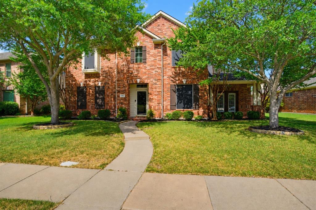 Welcome to this two-story brick home framed by mature trees and a landscaped front yard, overlooking a neighborhood park. Inside, the light and neutral interior features wood-look plank flooring throughout much of the main living areas, arched openings, decorative trim, and a mix of recessed and updated lighting. The kitchen offers white cabinetry, granite countertops, stainless steel appliances, a geometric tile backsplash, and a breakfast bar with a sink that opens to the adjoining living area. Nearby, the main living area features a white brick fireplace with a substantial mantel, a shiplap accent wall, and multiple windows looking out to the backyard and pool. A convenient half bath and separate laundry room with built-in shelving add functionality downstairs. The primary bath includes separate vanities, a soaking tub, separate glass-enclosed shower, and a walk-in closet with built-in shelving and hanging space. Upstairs, additional carpeted bedrooms are joined by a full bath, while a large open flex space provides additional living or game-room potential and includes a built-in desk area. Outside, the fenced backyard centers around an in-ground pool with a rock feature and surrounding patio space. A covered patio with a ceiling fan provides additional space for outdoor seating and entertaining.