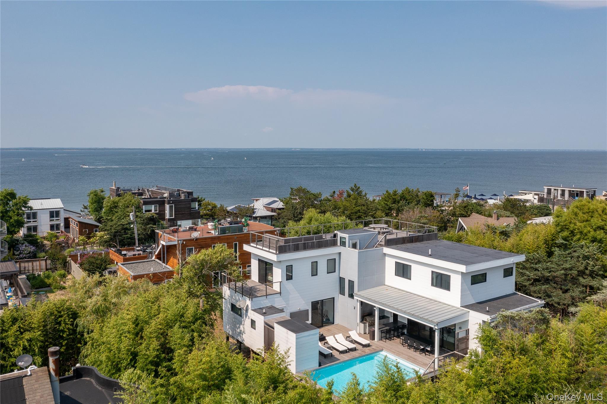 Experience coastal luxury at its finest with this exceptional Seaview retreat. Thoughtfully designed with over 3,650 square feet of living space, this stunning modern home offers 5 spacious bedrooms, 4 full bathrooms and 2 half baths, creating the perfect setting for effortless beach living and entertaining.The sun-filled main level welcomes you with soaring 10.5-foot ceilings and an expansive great room anchored by a fireplace and custom built-ins. A chef’s kitchen, appointed with quartz countertops and premium appliances, opens seamlessly to the living and dining areas. While a cozy den, large screened in porch, guest suite, powder room & laundry room complete the first floor.  A dramatic 16-foot wall of sliding glass doors extends the living space outdoors to an expansive mahogany deck, where a shaded pergola, fully equipped outdoor kitchen with built-in grill, and a heated 15’ x 25’ saltwater pool create the ultimate backyard oasis. Outdoor shower, cabana storage and half bathroom complete the main deck area. Upstairs, four beautifully appointed bedroom suites provide comfort and privacy, including a spectacular primary suite featuring bay views, a sitting area, spacious his & hers walk-in closets, spa-inspired bath with soaking tub, oversized shower and private water closet. End your day on the rooftop deck, taking in the sunset over the breathtaking 360-degree views of both the Great South Bay and Atlantic Ocean. Only minutes to Seaview’s beloved community amenities; playground, basketball courts, lifeguarded bay beach, kiddie pool, and tennis courts, this remarkable home offers an unparalleled Fire Island lifestyle. A rare opportunity to own one of Seaview’s finest homes. Shown by appointment only.