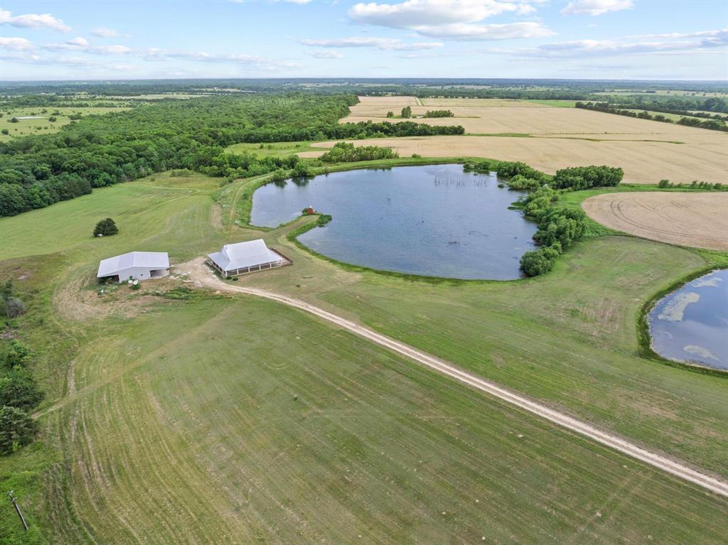 Escape the ordinary on this stunning and secluded 135+ acre retreat just 60 miles from Dallas. Tucked off FM 824 in Lamar County, this rare property offers the perfect blend of privacy, recreation, and rural charm — with easy access to Bonham, Paris, and the wider DFW metroplex.
A 10-acre stocked pond teeming with bass, perch, crappie, and catfish is the crown jewel of the land, offering anglers and nature lovers endless delight. Migratory ducks frequent the water, and deer roam the fields — making this a hunter’s haven. A second pond, a seasonal creek cutting through the back of the property, and fields, some of which are currently in wheat harvest, round out the diverse landscape. The 2,000 sq ft, 2-bed, 2-bath, 2-living area home was built in 2015, is nicely appointed with hardwood floors, granite counter tops, a WOLF gas cooktop and foam insulation in the attic making it energy efficient in those hot summer days. This home was designed for enjoying the outdoors — with a wide 360° wraparound porch perfect for soaking in both sunrises and sunsets over the big Texas sky. Towering pecan trees frame the entrance, and a 1,500 sq ft workshop with loft provides ample space for equipment, hay, or hobbies. The workshop also includes a 500 sq ft, climate-controlled room with kitchenette. There’s an underground storm cellar, butane powered Generac generator that can power the home and workshop, 500 gal. diesel tank and an RV pad with hookups, ready for guests or extended stays. Fully fenced and rich with potential, this land offers opportunity for livestock, equestrian use, or your own peaceful getaway. Whether you’re seeking a weekend retreat, a secluded homestead, or a sportsman’s paradise, this property delivers freedom, beauty, and breathing room — just far enough to feel away from it all.