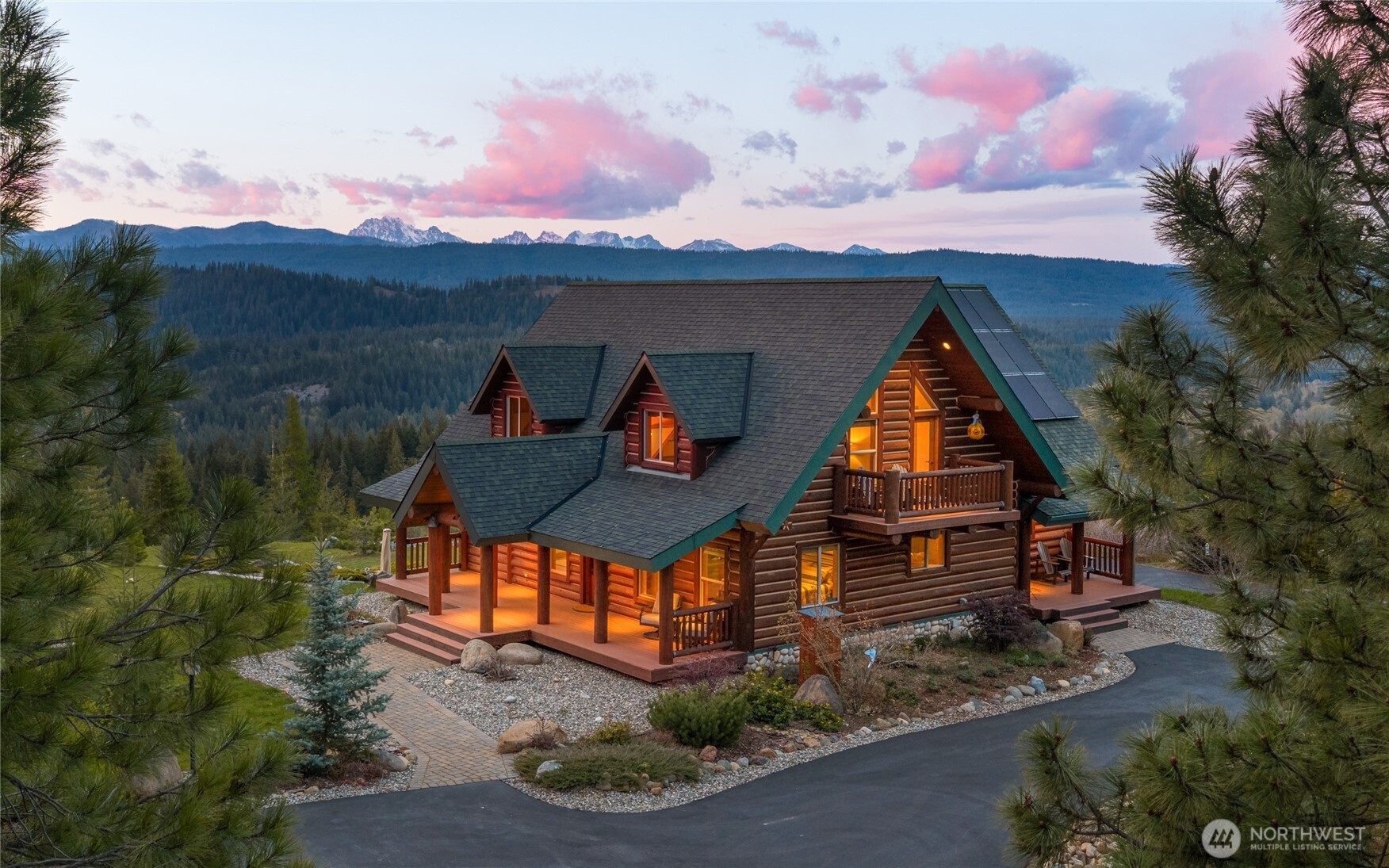 A striking mountain retreat framed by panoramic views, with warm interior lighting highlighting the home’s log construction and expansive covered porches.