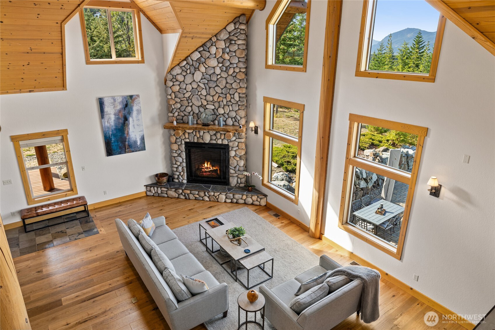 An upper-level view captures the soaring great room, floor-to-ceiling river rock fireplace, and dramatic wall of windows.