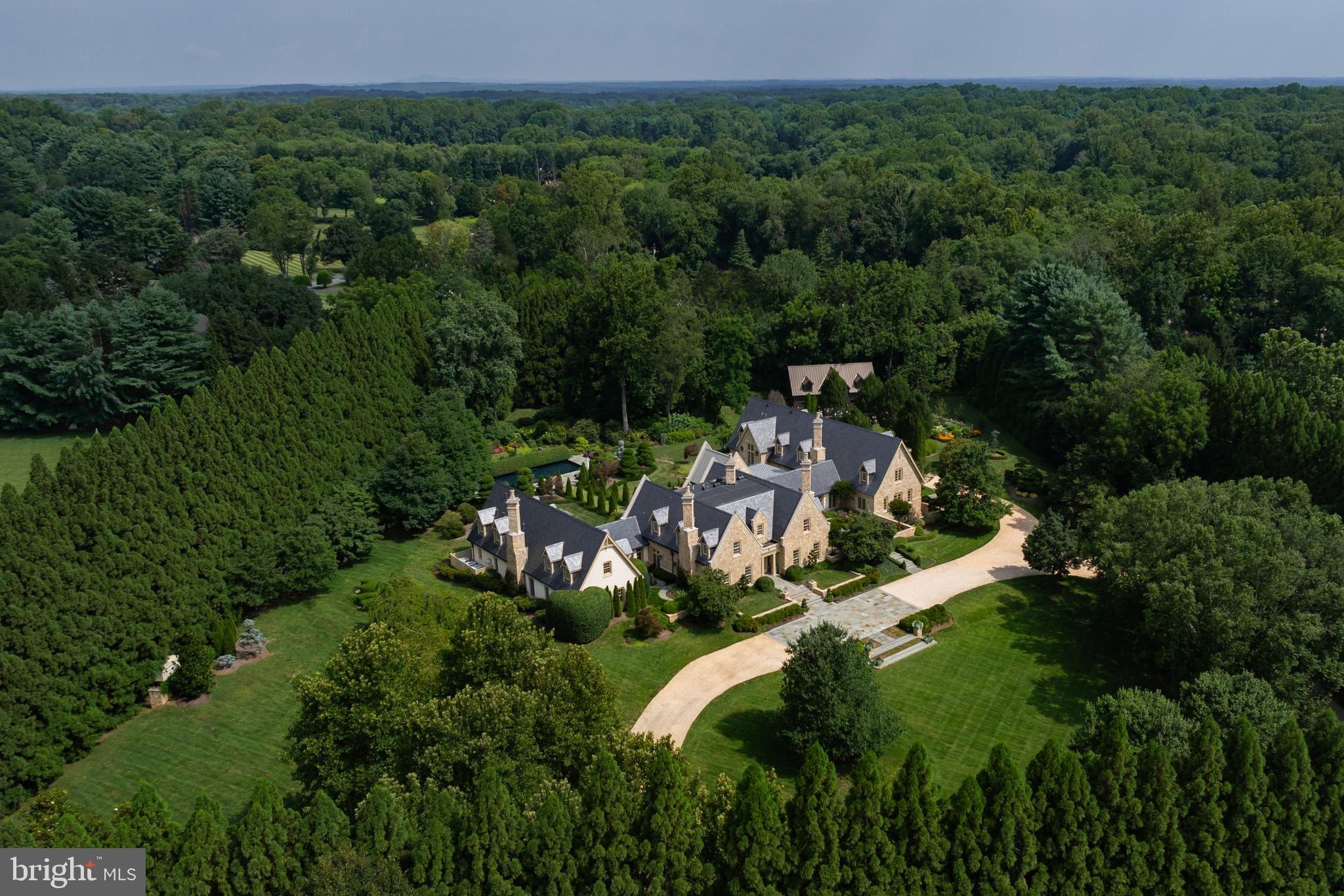 Set among the rolling hills of Great Falls, Alderley Manor is a five-acre estate inspired by the architecture and landscapes of the English Cotswolds, where light-stone manors rise above open expanses and long vistas. The residence was a collaboration between David Cooper of ACG Architects and BOWA Construction, with further enhancements by Hanlon Design Build. Spanning more than 20,000 square feet, the estate combines architectural integrity, exceptional craftsmanship, and highly considered amenities on a remarkable scale.A pea stone driveway winds through the grounds to a central flagstone motor court, introducing a residence defined by imported Cotswold limestone, a slate roof with metal seams, copper gutters, and finely finished woodwork. Classic symmetry, steep gables, and towering chimneys establish its distinctly English character, while rich mahogany frames every exterior window and door. Inside, reclaimed antique oak floors, hand-finished millwork, exquisite moldings, hand-carved details, custom built-ins, and oversized fireplace mantels with marble hearths carry the same level of craftsmanship throughout. Formal and informal rooms are generously proportioned yet inviting, with a layout that maintains symmetry while allowing for a natural flow between spaces.The main level is organized around a light-filled gallery connecting the east and west wings. Formal rooms include a moody library with deep green built-ins and bold ceiling wallpaper, while the informal spaces center around the chef's kitchen, appointed with twin islands, furniture-grade cabinetry, a Lacanche range, and artisanal tilework. The adjacent breakfast room features exposed beams, a stone accent wall, and multiple French doors opening to the grounds. The west wing is devoted to the primary suite, comprising a private garden, sitting room, dual dressing wings, and two luxurious baths. Two offices, including one with a vaulted beamed ceiling, complete the level.Upstairs, four bedrooms each have a distinct design sensibility and are served by Lagos Azul limestone baths with Waterworks fixtures. The lower level extends the home's entertaining and wellness spaces, with a transverse gallery leading to a professional cinema, climate-controlled wine cellar, and fitness center with spa bath, steam shower, and cold plunge. Billiards and art studio spaces provide additional areas for recreation and creative pursuits.The estate's infrastructure is equally considered. Commercial-grade systems include a self-sustaining well and septic system, whole-house generator, Lutron lighting, smart HVAC, and comprehensive security, providing seamless operation and independence commensurate with the scale of the property.Outside, the gardens are a defining element of Alderley Manor. An investment exceeding eight figures in landscaping and horticulture has created a highly composed landscape of manicured gardens, mature trees, and seasonal blooms. The grounds are fully irrigated and softly illuminated by custom landscape lighting, with hundreds of concealed fixtures bringing the property to life after dark. A heated lap pool with retractable cover, pergola-covered dining terrace, and newly installed sports court provide spaces for recreation, entertaining, and relaxation.For the automotive enthusiast, a modern farmhouse-style garage offers three bays, two lifts, a mezzanine lounge, and cigar bar. Despite its privacy, Alderley Manor is less than a mile from Great Falls Village and minutes from Tysons Corner, the Reston Tech Corridor, and Washington, D.C.With its rare combination of scale, architectural integrity, craftsmanship, infrastructure, and landscape, Alderley Manor stands among the region's most distinguished private estates.