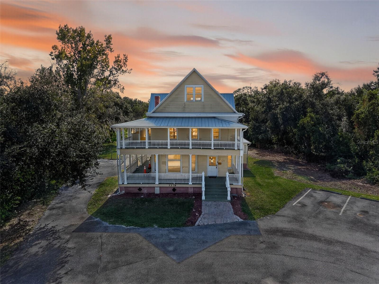 Welcome to an exceptional commercial opportunity at 1218 W Broad St in Groveland. This distinctive two-story property, rumored to be a historic structure, offers a generous footprint on a sizable lot with ample parking, making it suitable for a variety of office or retail uses. The exterior presents a classic facade with a welcoming full-length porch on both levels, featuring white exposed rafters, wood siding painted a light yellow, and large surrounding trees that offer shade. Inside, the property showcases beautiful original wood details, including a grand staircase with rich wood banisters and newel posts, and preserved original hardwood floors throughout the main rooms. The first level features expansive, well-lit spaces, including a room with a charming wood-framed brick fireplace, and another large area with multiple bay windows and an exterior exit, all featuring high ceilings and thick white baseboards. An intricately carved wooden archway adds historical character to the main floor. The upper level includes several bright rooms with contemporary light gray walls and original hardwood floors, providing flexible space for offices or other needs. One room features three large windows overlooking the grounds and a tin roof below. Another area on the upper level is an enclosed porch with wrap-around windows, a slatted ceiling, and light yellow horizontal wood paneling, offering a pleasant outdoor-like space. The property also includes a functional kitchen space with tile floors and wooden cabinetry, as well as a room with a standing shower stall, suggesting potential for various commercial layouts. This building offers a unique blend of historic charm and practical space in a prime location.