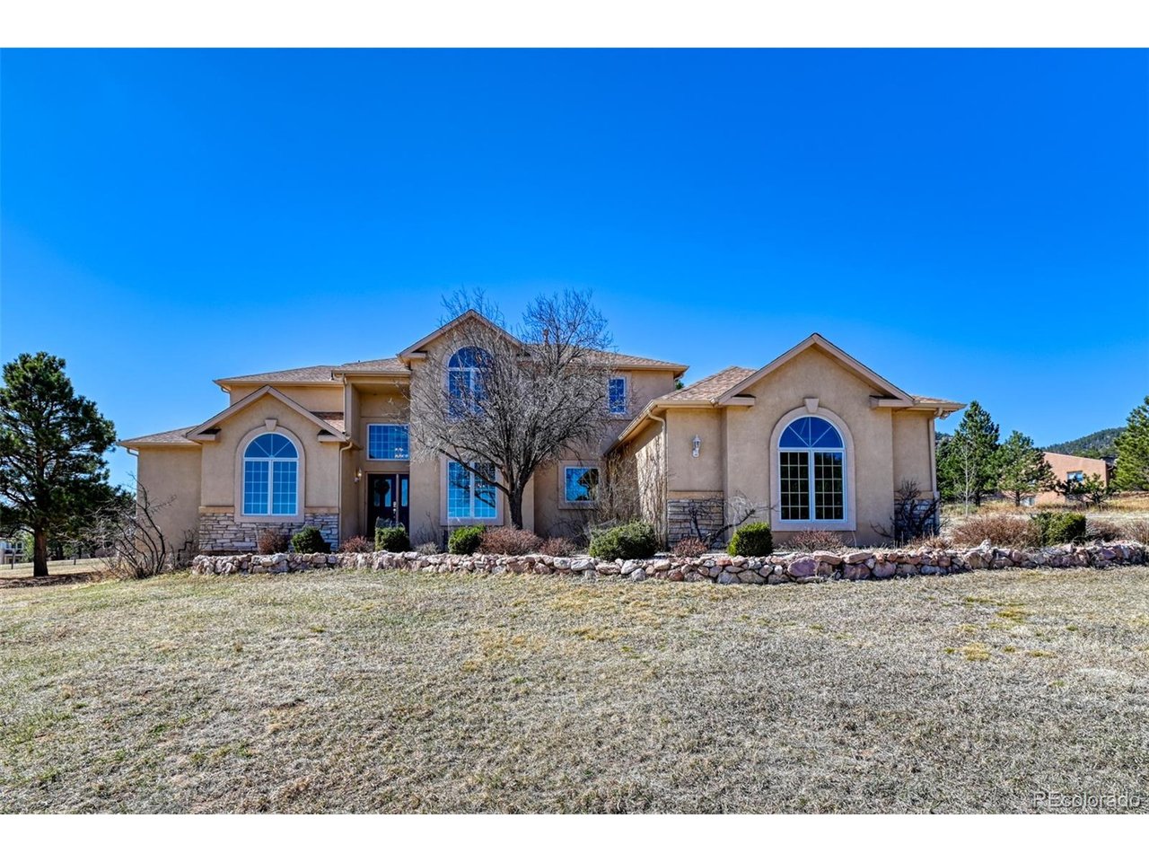 Beautiful custom stucco and stone two-story home with no covenants offering 4,826 sq ft of living space on a serene 2.28-acre lot filled with mature ponderosa pines & views of Elephant Rock out the front yard. Located in the highly desirable Lewis-Palmer School District and ideally situated between downtown Palmer Lake and Monument with convenient access to restaurants, shopping, and everyday amenities. No HOA. This 4-bedroom, 4-bathroom home features a spacious and thoughtfully designed floor plan with a 3-car garage. The dramatic great room showcases soaring vaulted ceilings and a stunning floor-to-ceiling stone gas fireplace, creating a warm and inviting centerpiece for the home. The main-level primary suite provides comfort and privacy, while three additional bedrooms and two bathrooms are located upstairs. The main floor also includes a dedicated office with French doors and built-in shelving, plus a formal dining room perfect for entertaining. The kitchen features a large island, abundant cabinetry, and generous counter space, opening to a back deck ideal for outdoor grilling and relaxing among the trees. Hardwood floors and Pella windows with built-in blinds throughout add both beauty and convenience. Additional highlights include a well-appointed laundry room with ample cabinetry, an unfinished basement with rough-in plumbing for a future bathroom, and a detached storage shed for lawn equipment and outdoor gear. One can add their own updated touches to this incredible home.