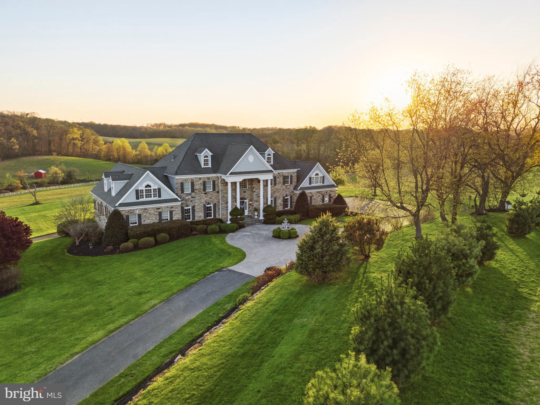 Set across approximately 115 extraordinary acres in the rolling countryside of White Hall, this landmark estate presents a rare opportunity to own a custom hand-built masterpiece by renowned builder Art Wood, whose name is synonymous with exceptional craftsmanship, architectural integrity, and enduring quality throughout Harford and Baltimore Counties. Privately sited amid panoramic rural vistas, the property offers an unmatched sense of scale, beauty, and permanence, with manicured grounds, extensive stone hardscaping, elegant retaining walls, covered patios, curated walkways, and a striking entry courtyard that creates an unforgettable arrival. Constructed in 2006, the main residence encompasses more than 10,000 square feet of finished living space across four finished levels, each thoughtfully designed to balance grandeur, comfort, and function. With 9 bedrooms, 5 full baths, and 3 half baths, the home is exceptionally appointed with an elevator, geothermal heating, a propane-powered whole-house generator, and a 3-car garage. Luxury spaces throughout include a private in-law apartment, theater room, game room, wet bar, library, fitness room, art room, and substantial storage, creating a residence equally suited for elegant entertaining, multigenerational living, and private retreat. A grand courtyard entry opens to a dramatic two-story foyer showcasing gleaming porcelain flooring, a sweeping dual curved Brazilian cherry staircase, retractable crystal chandelier, and an impressive overlook above. The soaring family room is anchored by a two-sided fireplace and framed by oversized windows that capture the surrounding countryside. At the heart of the home, the gourmet kitchen is richly appointed with granite countertops, Brazilian cherry flooring, a substantial center island, Viking gas range, Italian carved marble stove canopy, Italian tilework, wine storage, a walk-in pantry, and a sunlit dining area with French doors leading to the expansive elevated deck. The main-level primary suite offers a luxurious private retreat with soaring two-story ceilings, a fireplace, private deck access, two oversized walk-in closets with dressing areas and window seats, and a spa-inspired bath featuring a soaking tub, tile shower, dual vanities, separate water closets, and custom cherry cabinetry. Formal living and dining rooms, a mudroom, spacious laundry room, and additional refined gathering spaces further enhance the main level. Upper levels continue the home’s remarkable scale with multiple bedrooms, full baths, bonus spaces, exercise area, and storage, while the walkout lower level is designed for entertaining with a raised-hearth stone fireplace, theater with recliner seating, game area, wet bar, and private in-law quarters complete with a full kitchen and bath. Beyond the main residence, the estate includes a separate 3-bedroom, 2.5-bath Cape Cod tenant house built in 2007, offering exceptional flexibility for guests, extended family, or on-site management. Equestrian and agricultural amenities further distinguish the property and include a bank barn, 9-stall horse barn, 90x42 machinery shed, fenced pastures, stream, pond with island, wooded acreage, and productive tillable land. This is far more than a home. It is a legacy estate of rare caliber, hand-built by Art Wood and offering an extraordinary combination of craftsmanship, acreage, privacy, and presence in one of Harford County’s most scenic countryside settings.