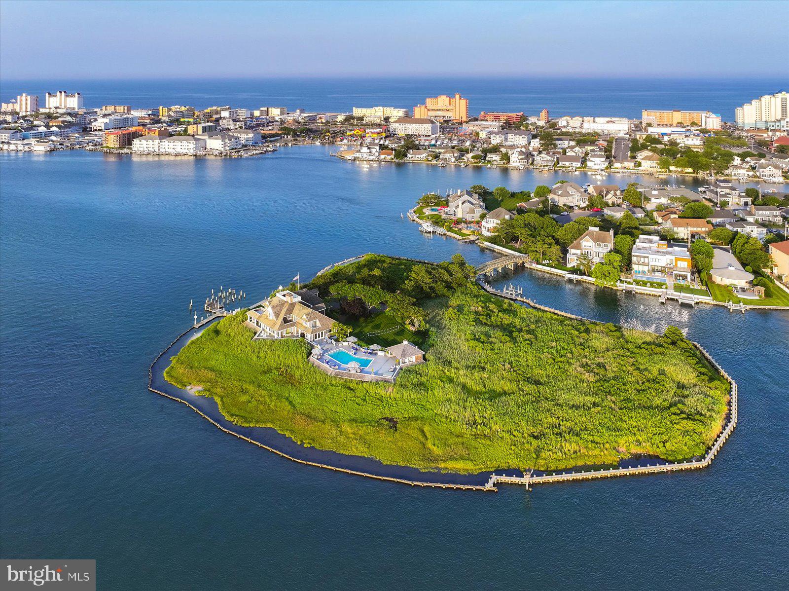 Goose Island at 1546 Teal Drive, Ocean City, Maryland is a truly unique offering — the bay’s only island estate. Located off 15th Street in downtown Ocean City, the property offers a remarkable balance of seclusion and accessibility,  3 blocks to the Boardwalk and beach, minutes to the Ocean City Inlet and Assateague Island by boat, and a block from Harbour Island, home of the world-renowned White Marlin Open. Hidden behind a brick wall extending along the property line and accessed through a 6-foot gate, the arrival experience continues across a private 120-foot vehicular bridge from Mallard Island. The island encompasses more than 3 acres of native marshlands and landscape with panoramic views of the open bay, the Rt. 50 Bridge to the south, the Rt. 90 Bridge to the north, and the rare combination of both breathtaking sunrises and sunsets across the water. Architecturally, the home is an iconic shingle-style residence. Inspired by Nantucket architecture, the residence underwent extensive exterior renovations and architectural enhancements under the direction of renowned Lyman Perry Architects Ltd. of Nantucket, Massachusetts, with Lyman Perry personally consulting on the estate. The approximately 5,000-square-foot home blends timeless design with exceptional craftsmanship, featuring custom woodworking, Carlisle wide-plank hand-finished pine flooring, new Andersen doors throughout, approximately 3,250 square feet of wraparound deck, and a resort atmosphere. Upon entering, a dramatic 24-foot atrium features an expansive 12-foot by 15-foot overhead skylight and a floor-to-ceiling 24-foot Coral Rock fireplace feature that anchors and connects the surrounding living spaces while filling the home with natural light. Designed to maximize open bay views throughout the home, the main living area features custom built-in cabinetry and entertainment center while flowing naturally into the separate dining and gathering spaces. The open-concept kitchen/family room floor plan creates an inviting living area designed for entertaining. Custom solid birch painted cabinetry by Premier Cabinetry of New Holland, Pennsylvania, includes a generous center island with a 5-foot by 8-foot teak countertop. A custom wood spiral staircase in the kitchen/family room provides secondary access to the upper level. The main level also includes a home office with built-in cabinetry and concealed pull-out file storage, along with an additional guest bedroom and full bath. Upstairs, an open bridge hallway overlooks the atrium and Coral Rock feature while separating the primary suite from two guest bedroom suites. The four-room primary suite consists of a bedroom retreat with access to a bayfront deck, a combined sitting, entertainment, and dressing room with vaulted ceilings, custom cherry wood built-ins and four closets, an en-suite bath with double vanity and dressing table, and a second dressing room featuring vaulted ceilings, four double-door deep closets, and an arched window with window seat dividing two built-in tall chests totaling 30 drawers. Two additional upstairs bedrooms each feature en-suite baths and private decks. Outdoors, the property transforms into an island retreat with a pool and spa by Sea Glass Pools, expansive decking, a large entertaining gazebo bar with cabinetry and quartz countertops, a full bath with shower, and protected docking facilities with parallel docking along approximately 1,600 feet of bulkhead, a 10,000-pound 2024 boat lift, and a fish cleaning station. Significant recent investments include bulkhead improvements, reinforced helical pilings, extensive bridge renovations, active security and surveillance systems, and meticulously maintained structural and mechanical systems. Surrounded by abundant birdlife including osprey, great blue herons, great white egrets, snowy egrets, mallards, and bald eagles, along with endless open-water vistas, Goose Island is a distinguished estate defined by timeless elegance.