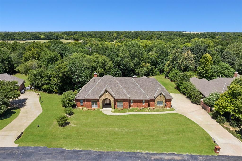 Truly beautiful home nestled on a private 1.11 acre wooded greenbelt lot in the desirable gated Deer Creek Hills addition! This exceptional home showcases gorgeous hardwood floors throughout, custom finishes, and a thoughtfully designed floor plan that blends elegance with everyday comfort. Arrive through the side-entry 3-car garage, complete with a storm shelter for peace of mind. As you make your way to the front entrance, a charming barrel wood ceiling and arched front door welcomes you. Step inside to an inviting open-concept layout. A formal dining room sits on one side of the entry, while a versatile formal living room on the other can easily serve as an office, sitting room, or flex space. Straight ahead, expansive windows frame breathtaking views of the wooded backyard, drawing your eyes outdoors. The stacked stone fireplace serves as the centerpiece of the living area, creating a warm gathering space. The open kitchen is designed for entertaining, featuring beautiful granite countertops, rich stained wood cabinetry with glass-front accents, built-in appliances, a refrigerator, and an oversized island perfect for preparing meals or gatherings. The private primary suite is a true retreat, complete with its own fireplace and double doors leading directly to the covered back patio. The spa-inspired ensuite offers dual vanities, a large soaking tub, a tiled walk-in shower, and an oversized walk-in closet with built-in drawers. Two additional bedrooms each enjoy direct access to a shared bathroom with a tub/shower combination, generous vanity, and ample storage. The spacious laundry room includes a utility sink, extra counter space, and cabinetry. Step outside to the expansive covered patio overlooking the peaceful, tree-filled backyard. With plenty of room to add a pool or create your own outdoor oasis, the possibilities are endless. This beautifully maintained Deer Creek home offers privacy, space, and timeless style all at an exceptional value.