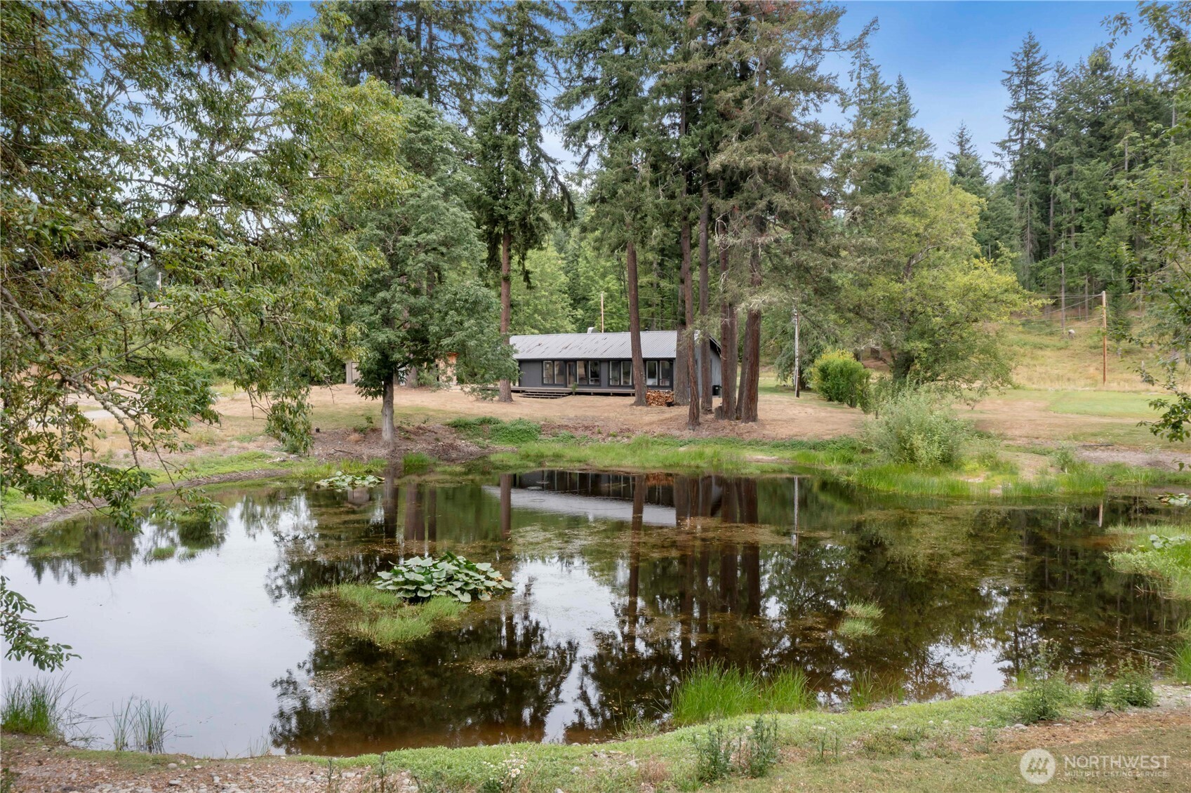 Escape to your own private retreat on 5 fully fenced acres! This serene property features a charming 2-bedroom cabin with a huge wraparound deck overlooking a tranquil pond—perfect for watching wildlife wander by. Inside, enjoy an open, light-filled living space with large windows that bring nature in, a sleek ductless mini-split for year-round comfort, stylish industrial-style plumbing, and a unique sliding barn door. The oversized detached 2-car garage has its own 220-amp meter—ideal for a workshop or hobbyist—and there's additional covered parking for toys or equipment. With a durable metal roof, no HOA, and total privacy, this is country living with modern flair!