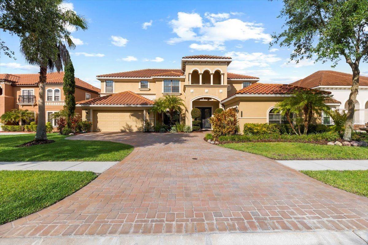 Sophistication, architectural elegance, and refined living converge in this former model home—an ICI Teramo design, entered into the 2013 Parade of Homes and showcasing exceptional craftsmanship and elevated finishes. Located within the exclusive, guard-gated Grand Hampton community, this 5-bedroom, 3.5-bath, 3-car garage estate is a Mediterranean-inspired masterpiece with no rear neighbors and serene pond views epitomizing Florida luxury living. Upon arrival, the curb appeal impresses—professionally manicured landscaping, timeless tile roof, custom paver driveway, and tropical palms frame the entry. A charming paved seating area welcomes guests in true resort style. Step through grand wood-framed glass double doors into an interior of meticulous design. The formal dining room, left of the foyer, features arched ceilings, architectural columns, and timeless sophistication. To the right, a private office with rich wood floors, crown molding, and a modern light fixture creates a refined workspace. The first-floor primary suite is a sanctuary with arched ceilings, decorative columns, warm wood floors, upgraded lighting, and a beautiful chandelier. A tranquil sitting area opens to the lanai with peaceful views. The expansive custom walk-in closet is thoughtfully designed. The en-suite bath boasts dual vanities, a garden tub with chandelier, and a walk-in shower —offering spa-level comfort. The main living area blends luxury and livability with dramatic marble wall paneling, tray ceilings, recessed lighting, and a designer chandelier. Panoramic three-panel glass sliders fully retract into the wall, creating seamless indoor-outdoor flow and framing stunning pond views. The gourmet kitchen features custom solid wood cabinetry with Certified KCMA 42-inch crown molding, granite countertops, under-cabinet and recessed lighting, new designer hardware, Stainless Steel Energy Star Appliances, and a spacious dining area. A vented pantry completes this chef’s dream. Also on the main level: a half bath, laundry room with upgraded cabinetry and utility sink, and an oversized 3-car garage with Smart Garage Opener. Ascend the staircase, highlighted by new iron balusters, to the second floor where four spacious bedrooms offer ample closets and natural light. A flexible media room/loft/gym features updated industrial hardwood flooring and opens to a rear balcony with tranquil pond views. Step outside to your private resort-style retreat. Custom paver decking surrounds a heated pool and spa finished with a non-slip, stain-resistant pebble-bottom surface. Enjoy three cascading water walls, multicolor lighting, a gas quick-connect for grilling, and Florida Glass privacy paneling. The fully screened lanai and fenced backyard feature elegant aluminum fencing, plus a child safety fence around the pool and a convenient dog door—perfect for safety, privacy, and entertaining. Notable upgrades include: 8-ft doors throughout both levels, HERS RESNET Certified, High-performance thermal-tinted windows and glass doors, Tankless hybrid water heater, Dual-zone HVAC systems, Programmable thermostats, Water-conserving fixtures, Security system, Newer pool heater. Grand Hampton offers resort-style amenities: clubhouse, fitness center, lap and resort pools, tennis courts, playgrounds, and walking trails. Ideally located near I-75 and I-275—minutes from AdventHealth, The Shops at Wiregrass, Tampa Premium Outlets and more. This home isn’t just luxurious—it’s a lifestyle.