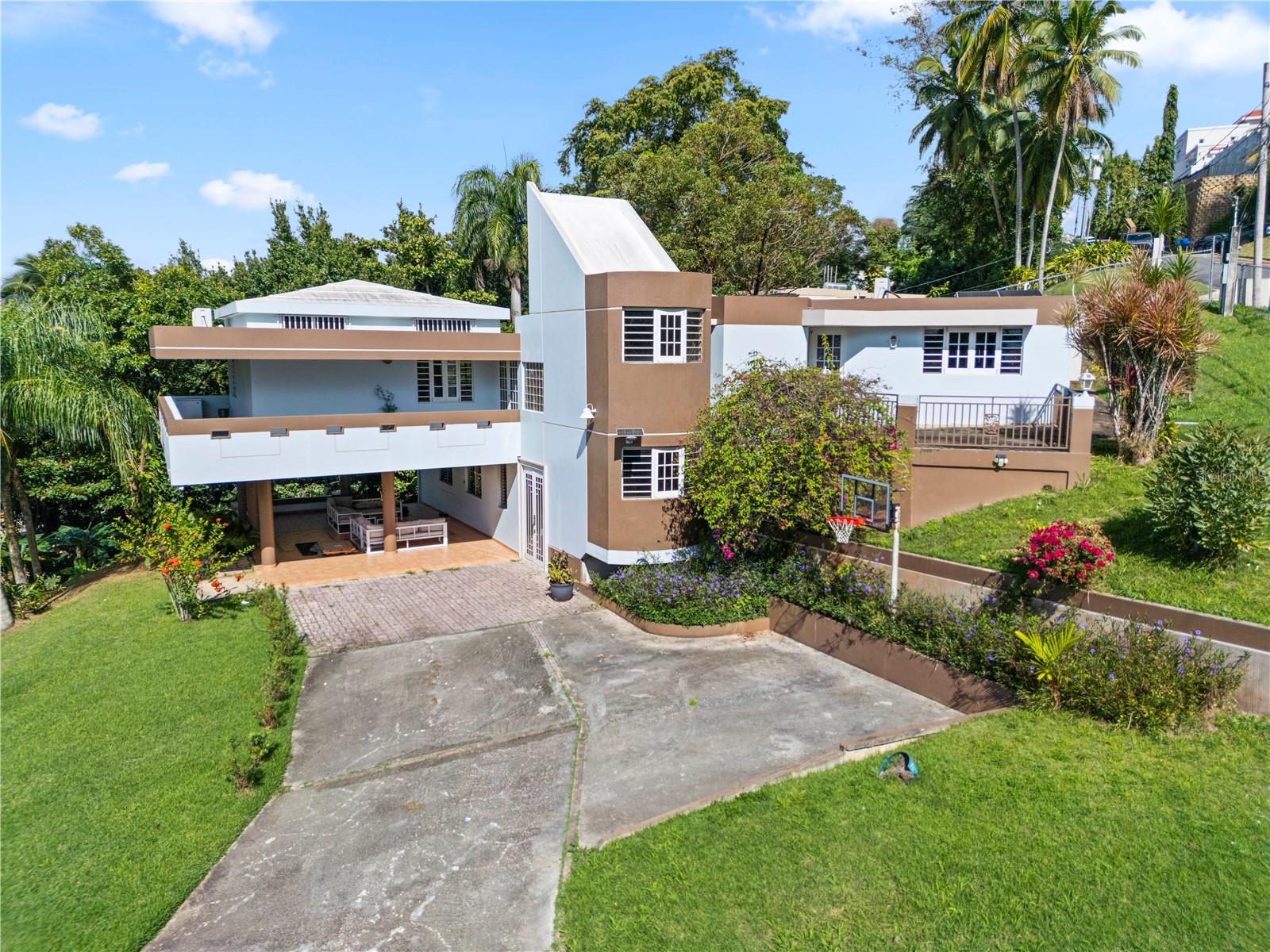 Located within the gated community of El Mirador de San Juan, this distinctive residence offers privacy, space, and a timeless architectural presence on an expansive half-acre lot surrounded by mature tropical landscaping. Avocado and plantain trees frame the property, creating a serene, garden-like setting that feels both established and tranquil.The home’s architecture blends clean lines with warm, modernist influences—defined by its strong geometric forms, elevated main living areas, and a wrap-around porch that extends the living room outdoors. Covered terraces and shaded outdoor spaces invite year-round indoor-outdoor living, ideal for entertaining or quiet moments surrounded by greenery.The main residence features 4 bedrooms and 3 bathrooms, with an updated kitchen anchoring the primary living area. On the first level, a private in-law apartment offers exceptional flexibility, complete with one bedroom, one bathroom, and a kitchenette—ideal for guests, extended family, or additional living arrangements.Designed with efficiency and sustainability in mind, the property is equipped with solar panels and two Tesla Powerwall batteries, providing energy resilience and long-term cost savings.Conveniently located just 3 minutes from Montehiedra Shopping Center, dining options, and daily conveniences, and only 4 minutes from Highway 52, this home offers a rare combination of privacy, accessibility, and space—set within one of San Juan’s most desirable residential enclaves.A unique opportunity to own a character-rich home with land, energy independence, and a prime San Juan location.