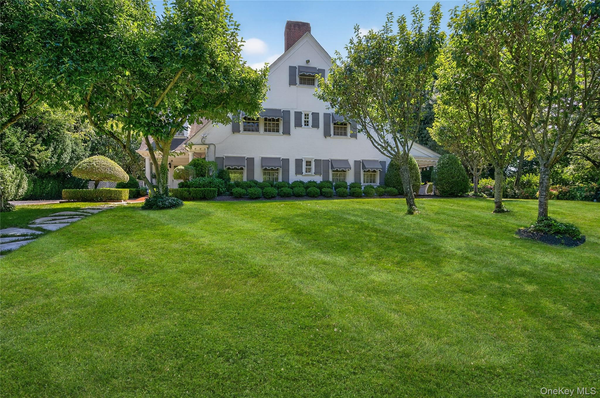 Discover timeless sophistication at 42 Clock Tower Lane, Old Westbury. Designed in 1891 by celebrated architect Stanford White, this distinguished residence was once part of the historic Morgan Estate and rests on more than 3 acres at one of Long Island’s highest points. Offering 5 bedrooms and 4.5 baths, the home is defined by soaring 10+ foot ceilings, intricate millwork, and eight fireplaces that exude warmth and character. Expansive formal rooms create an elegant yet inviting setting for both intimate gatherings and grand entertaining.
At the heart of the home is a gourmet eat-in kitchen, thoughtfully designed with premium finishes and sliding glass doors that open to a covered patio for seamless indoor-outdoor living. A mudroom off the kitchen and driveway features a first-floor washer/dryer, while a second washer/dryer is located in the basement for added convenience. Modern comforts include central air, a whole-house generator, security system, handicap accessibility, and a 2-car garage. Outdoors, the park-like grounds showcase perennial gardens, an estate-size pool with granite patio, and a charming pool house complete with kitchenette, changing room, full bath, and lounge area. A private tennis court completes the resort-like setting. Located within the award-winning Jericho School District, this property is a rare opportunity to enjoy the perfect balance of historic architecture and modern luxury living.