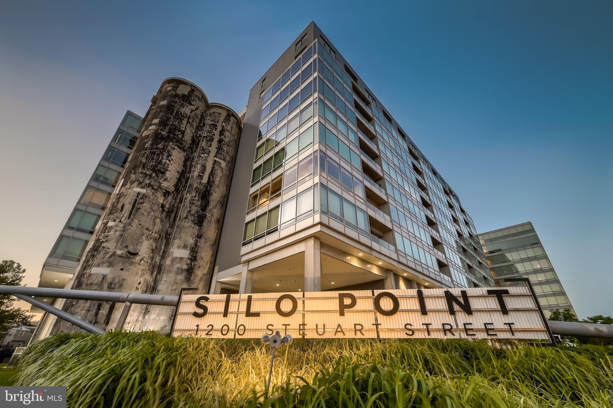 Thoughtfully updated and beautifully appointed, this spacious corner one-bedroom, one-and-a-half-bath residence at Silo Point blends modern design with the building’s iconic industrial character. An expansive open floor plan, wide-plank luxury vinyl flooring, and extensive track and directional lighting create a sophisticated yet comfortable setting for everyday living and entertaining.A gracious entry, complete with an oversized walk-in closet, opens to the generous living and dining area, where a statement chandelier defines the dining space. The open-concept kitchen is anchored by a large island with pendant lighting and complemented by sleek cabinetry, stainless steel appliances, under-cabinet lighting, and abundant storage, seamlessly connecting the kitchen with the surrounding living spaces. Just beyond, an expansive private balcony extends the home outdoors and features a ceiling fan for added comfort. The generously proportioned bedroom and dressing area offer a private retreat with new carpeting, custom built-in bookcases, upgraded lighting, a large wardrobe, and additional closet space. The en-suite bath features a double-bowl stone vanity, soaking tub, and separate walk-in shower. A powder room provides added convenience for guests, while the oversized laundry room offers additional storage and a full-size washer and dryer. One deeded garage parking space, conveniently located near the residence, is included. Experience the coveted Silo Point lifestyle with an impressive collection of full-service amenities, including a 24-hour front desk attendant, on-site management, a fitness center with yoga studio and treatment room, boardroom, game room, EV charging, and the spectacular 19th-floor Sky Lounge with sweeping harbor and city views. Ideally situated in the heart of Locust Point, residents enjoy convenient access to waterfront parks, restaurants, neighborhood favorites, the water taxi, and I-95. Distinctive architecture, generous living spaces, and an exceptional waterfront location define the Silo Point lifestyle.