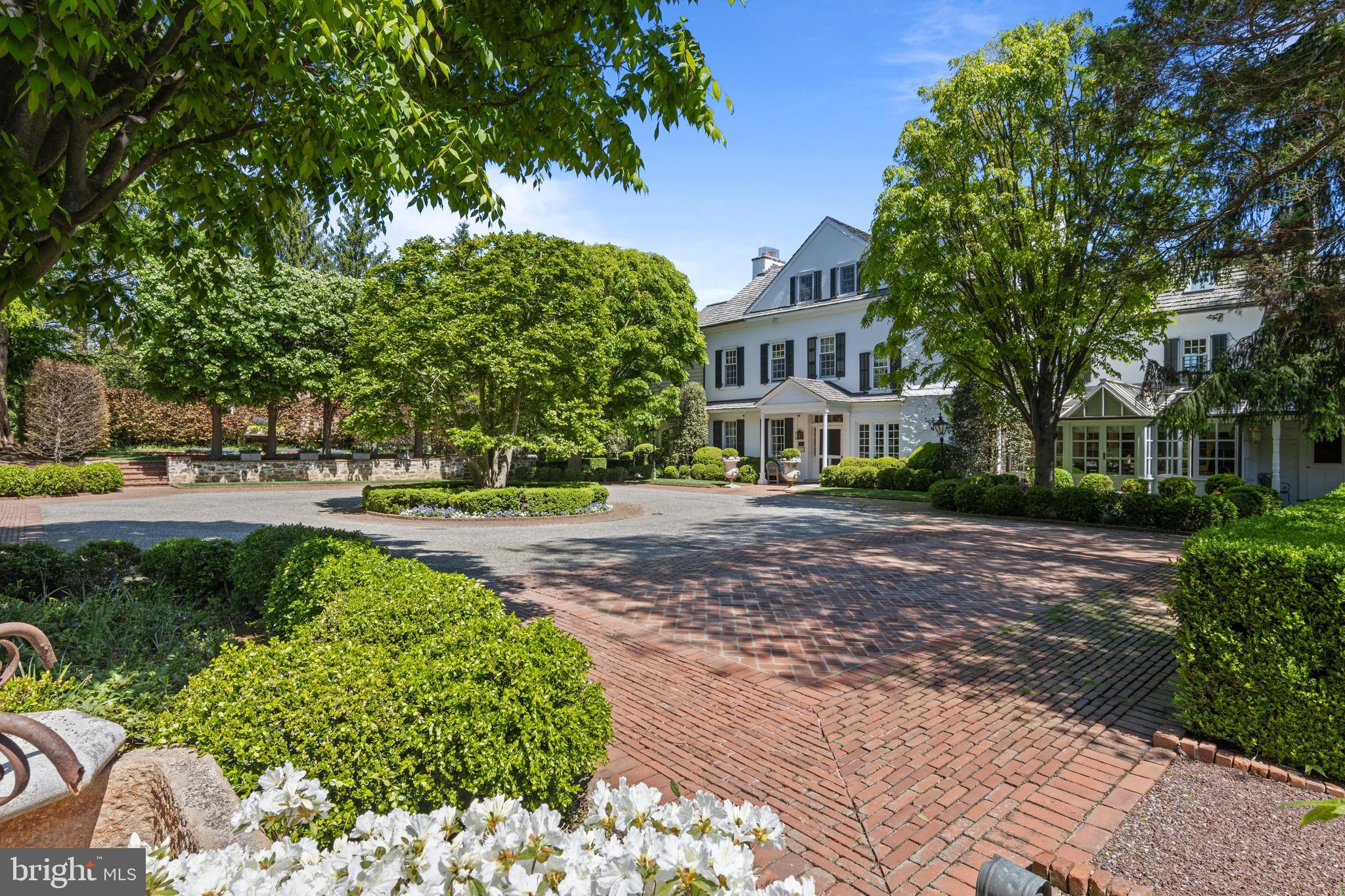 One of only six residences along a private Gladwyne lane, 1320 Huntsman Lane is a quietly masterful composition—where architecture and landscape are conceived as one continuous experience, and where restraint, proportion, and craftsmanship define its character.A painted white stone farmhouse anchors the end of a wide gravel court, the approach unfolding in glimpses. A beech lawn, enclosed by mature trees and patterned with a restrained formal garden, introduces the first of many outdoor rooms. Elsewhere, a stone wall with an arched opening leads to a more intimate enclosure and a separate structure suited for a studio, gym, or private office—each element placed with intention rather than display.Inside, the home honors its origins while elevating them. A straight-through foyer with richly stained original floors opens to terraced gardens beyond. Principal rooms balance refinement with ease: a living room extends to a screened garden space overlooking a formal walk of towering trees and cutting gardens; a fireside library offers quiet contrast; and a dining room opens seamlessly to a bluestone terrace. A glass-wrapped solarium adapts effortlessly to daily life, always oriented toward the landscape.The kitchen and adjoining spaces continue this dialogue. Substantial materials—wide-planked floors, marble surfaces, professional appliances—are paired with uninterrupted views outward. A glass-enclosed breakfast room opens to a wisteria-covered pergola with a fireplace, creating a space defined as much by nature as by structure. Nearby, a large family room offers a more relaxed counterpoint, with a detailed wood ceiling, wraparound windows evoking a treehouse-like setting, and a fireplace with concealed media—equally suited for gathering or retreat.Upstairs, an arched stair window frames the symmetry of the gardens below. The primary suite is warm and composed, with thoughtfully divided baths, tailored dressing spaces, and views to the cutting gardens. Additional bedrooms are generous and well-appointed, while a private guest suite and upper-level retreat provide flexibility for extended stays. A light-filled gym, defined by exposed stone and high ceilings, completes the interior.Here, the grounds are not ancillary; they are integral. A sequence of garden rooms unfolds along stone and brick paths that reveal rather than present. A grass tennis court, enclosed by intricate lattice fencing, is both refined and restrained, with a charming shed positioned along its center line. An allée of trees leads to the pool—rectangular, softened by a waterfall and balanced by curved seating—while two stone pavilions, seamlessly integrated, shelter dining and a waterfall-edge hot tub.A fire pit and a discreet clock tower with bath and changing space allow days to extend fully outdoors. Specimen plantings, antique garden elements, and thoughtfully placed lighting lend a sense of permanence, while centennial brick and stone paths ground the composition.At 1320 Huntsman Lane, beauty is not singular or immediate—it is cumulative, revealed gradually through movement, season, and time.