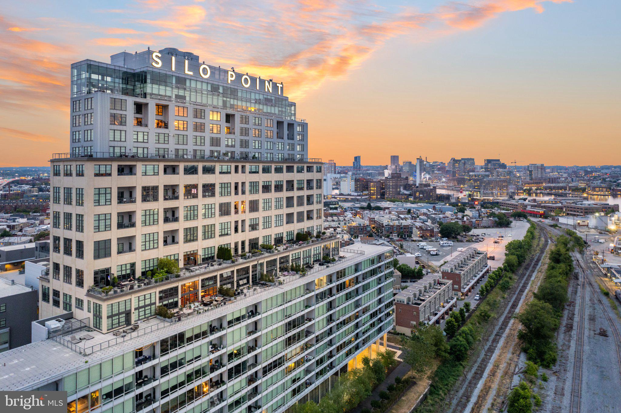 Set high on the 15th floor of Silo Point Tower, this exceptional corner residence showcases spectacular panoramic water and city views from coveted triple exposures to the east, west, and south. Sunrises and sunsets unfold through expansive windows, bathing the home in natural light throughout the day. Offering more than 2,200 square feet, this distinctive two-bedroom, two-and-a-half-bath condominium combines dramatic architectural scale with sophisticated, comfortable living. Soaring 14-foot ceilings and floor-to-ceiling windows create an extraordinary sense of volume, while exposed columns and ductwork add to the residence’s signature industrial aesthetic. Wood floors extend throughout the open, light-filled interiors, complemented by Lutron motorized blinds. Generous living and dining spaces provide an ideal setting for both relaxed daily living and effortless entertaining. At the heart of the home, the chef’s kitchen features a substantial granite island, tile backsplash, and custom cherry cabinetry accented with distinctive aluminum band detailing. The spacious primary suite serves as a serene private retreat, highlighted by floor-to-ceiling windows, custom closets, and a luxurious spa-inspired bath with dual vanity and an oversized glass-enclosed walk-in shower. A well-appointed second bedroom with its own en-suite bath offers flexibility for guests, a home office, or both. With its dramatic proportions, abundant natural light, exceptional views, and refined industrial character, this remarkable residence offers a rare combination of grandeur and warmth. Two premium garage parking spaces and a private storage unit convey. Experience the coveted Silo Point lifestyle with full-service amenities including 24-hour attendant, on-site management, a state-of-the-art fitness center with yoga studio and treatment room, boardroom, game room, and the spectacular 19th-floor Sky Lounge. Conveniently located with easy access to I-95 and the water taxi, and set in the heart of Locust Point—one of Baltimores most sought-after waterfront neighborhoods offering walkability to parks, dining, and vibrant city living.