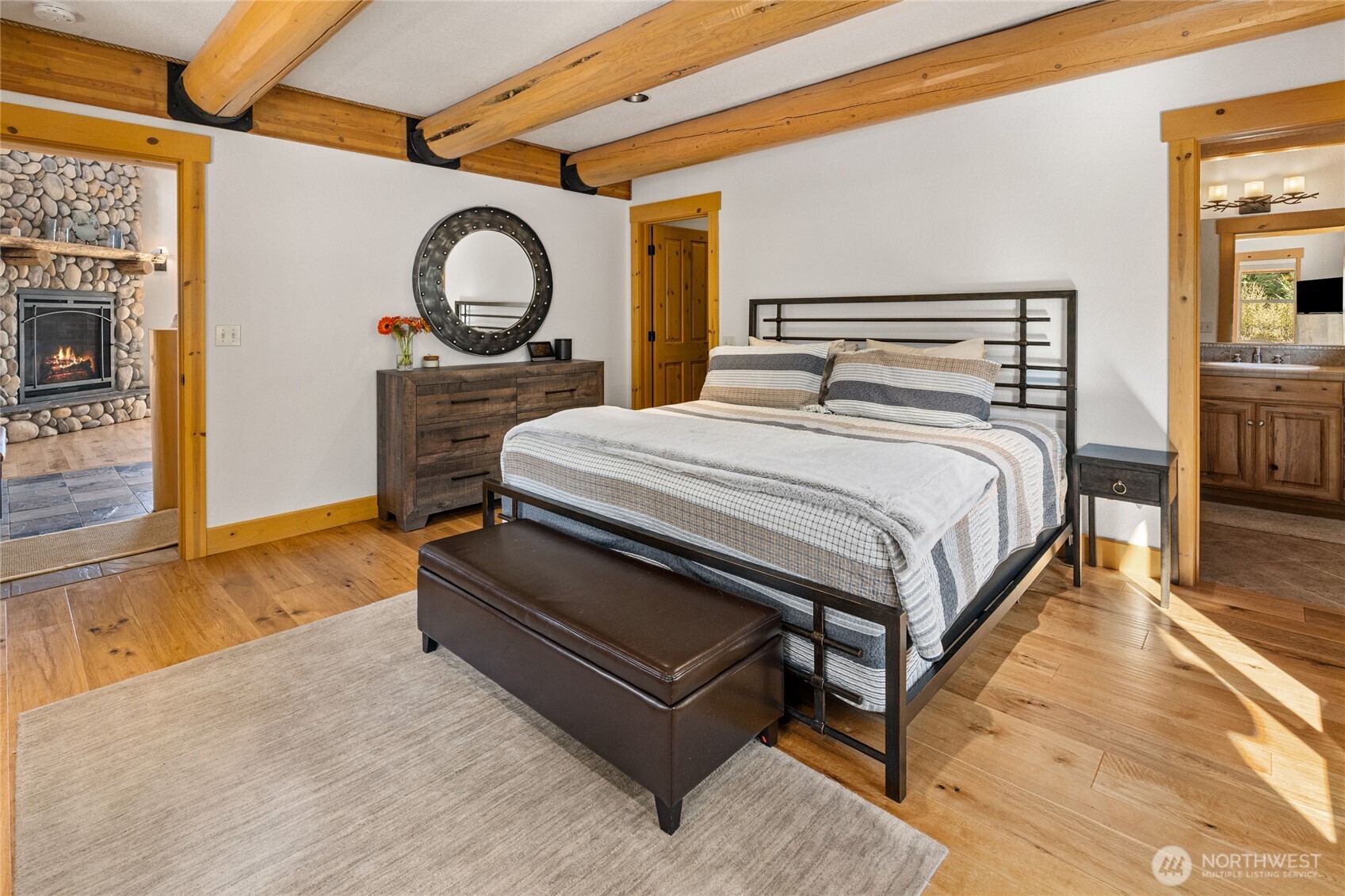 Main-level primary bedroom with hardwood floors, exposed log beams, warm wood details, and abundant natural light.