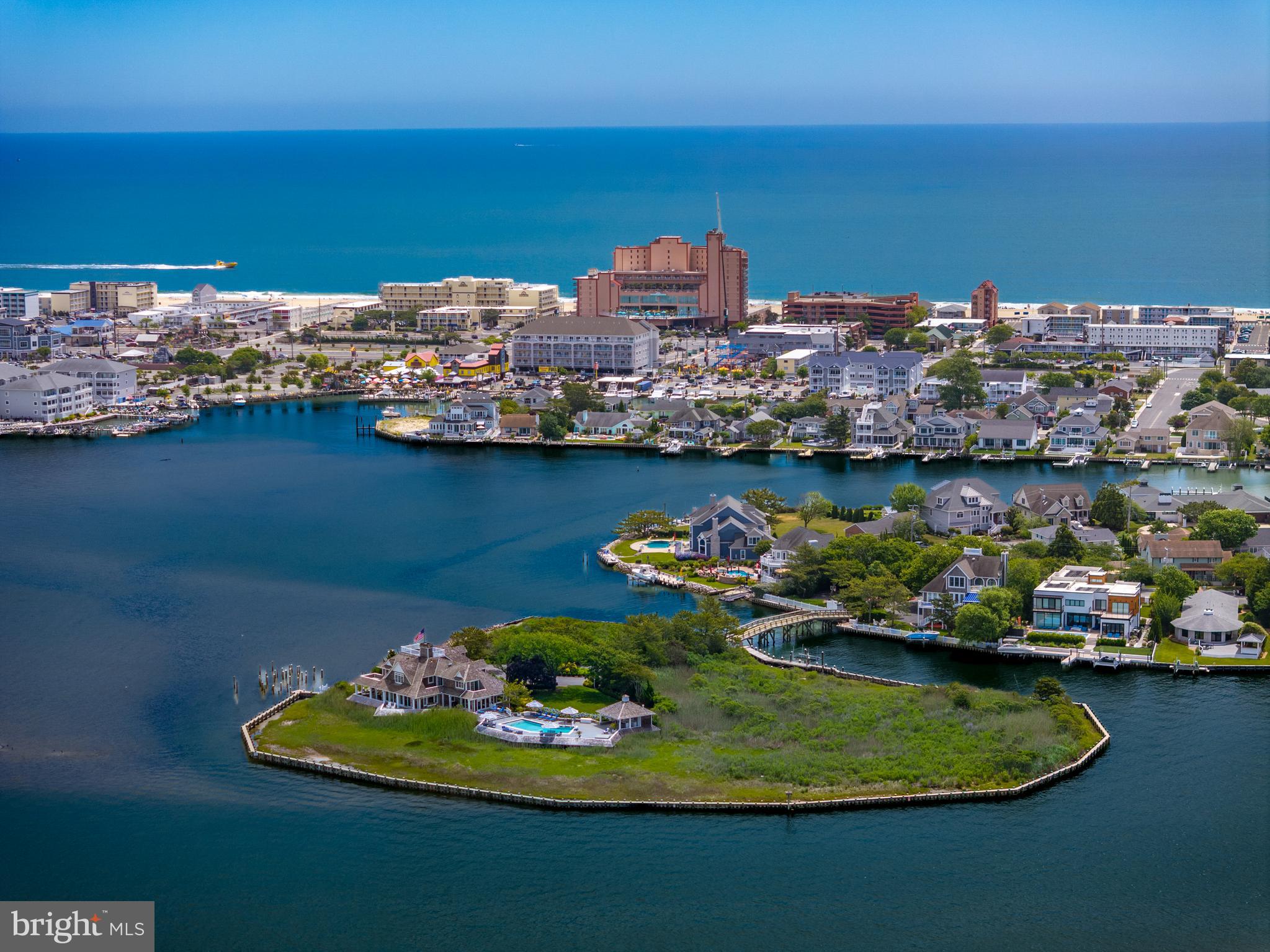 Private Island at 1546 Teal Drive, Ocean City, Maryland is a truly unique offering — the bay's only island estate. Included is an adjacent buildable waterfront lot just inside the entrance gates before crossing the private bridge. The improved .29-acre parcel features approximately 95 feet of bulkheaded shoreline with boardwalk, paved driveway, irrigation, and landscaping, offering flexibility for a future guest house or single-family residence, additional parking, or expanded waterfront enjoyment. Located off 15th Street in downtown Ocean City, the property offers seclusion and accessibility, 3 blocks to the Boardwalk and beach, minutes to the Ocean City Inlet and Assateague Island by boat, and a block from Harbor Island, home of the world-renowned White Marlin Open. Hidden behind a brick wall and accessed through a 6-foot gate, the arrival continues across a private 120-foot vehicular bridge from Mallard Island. The island encompasses more than 3 acres of native marshlands and coastal landscape with panoramic views of the open bay, the Rt. 50 Bridge to the south, Rt. 90 Bridge to the north, and the rare combination of breathtaking sunrises and sunsets across the water. Architecturally, the home is an iconic shingle-style residence inspired by Nantucket architecture, with exterior renovations and enhancements by renowned Lyman Perry Architects Ltd. of Nantucket, Massachusetts. The approximately 5,000-square-foot home features custom woodworking, Carlisle wide-plank hand-finished pine flooring, new Andersen doors throughout, approximately 3,250 square feet of wraparound decking, and expansive water views. Upon entering, a dramatic 24-foot atrium features a 12-foot by 15-foot overhead skylight and floor-to-ceiling Coral Rock fireplace feature connecting the surrounding living spaces. Designed to maximize open bay views throughout, the main living area features custom built-in cabinetry and flows into the separate dining and gathering spaces. The kitchen/family room creates an inviting area for entertaining, with custom solid birch painted cabinetry by Premier Cabinetry of New Holland, Pennsylvania, and a 5-foot by 8-foot teak island countertop. A custom wood spiral staircase provides secondary access between the main and upper levels. The main level also includes a home office with built-in cabinetry and pull-out file storage, plus an additional guest bedroom and full bath. Upstairs, an open bridge hallway overlooks the atrium while separating the primary suite from two guest bedroom suites. The four-room primary suite includes a bedroom retreat with bayfront deck, sitting/entertainment/dressing room with vaulted ceilings, custom cherry wood built-ins and four closets, en-suite bath with double vanity and dressing table, and a second dressing room with vaulted ceilings, four double-door deep closets, and an arched window with window seat dividing two built-in tall chests. Two additional bedrooms each feature en-suite baths and private decks. Outdoors, an island retreat features a pool and spa by Sea Glass Pools, expansive decking, gazebo bar with cabinetry and quartz countertops, full bath with shower, docking, 10,000-pound boat lift, and fish cleaning station. Significant recent investments include bulkhead improvements, reinforced helical pilings, bridge renovations, active security and surveillance systems, and maintained structural and mechanical systems. Surrounded by abundant birdlife and endless open-water vistas, this distinguished estate embodies timeless elegance.