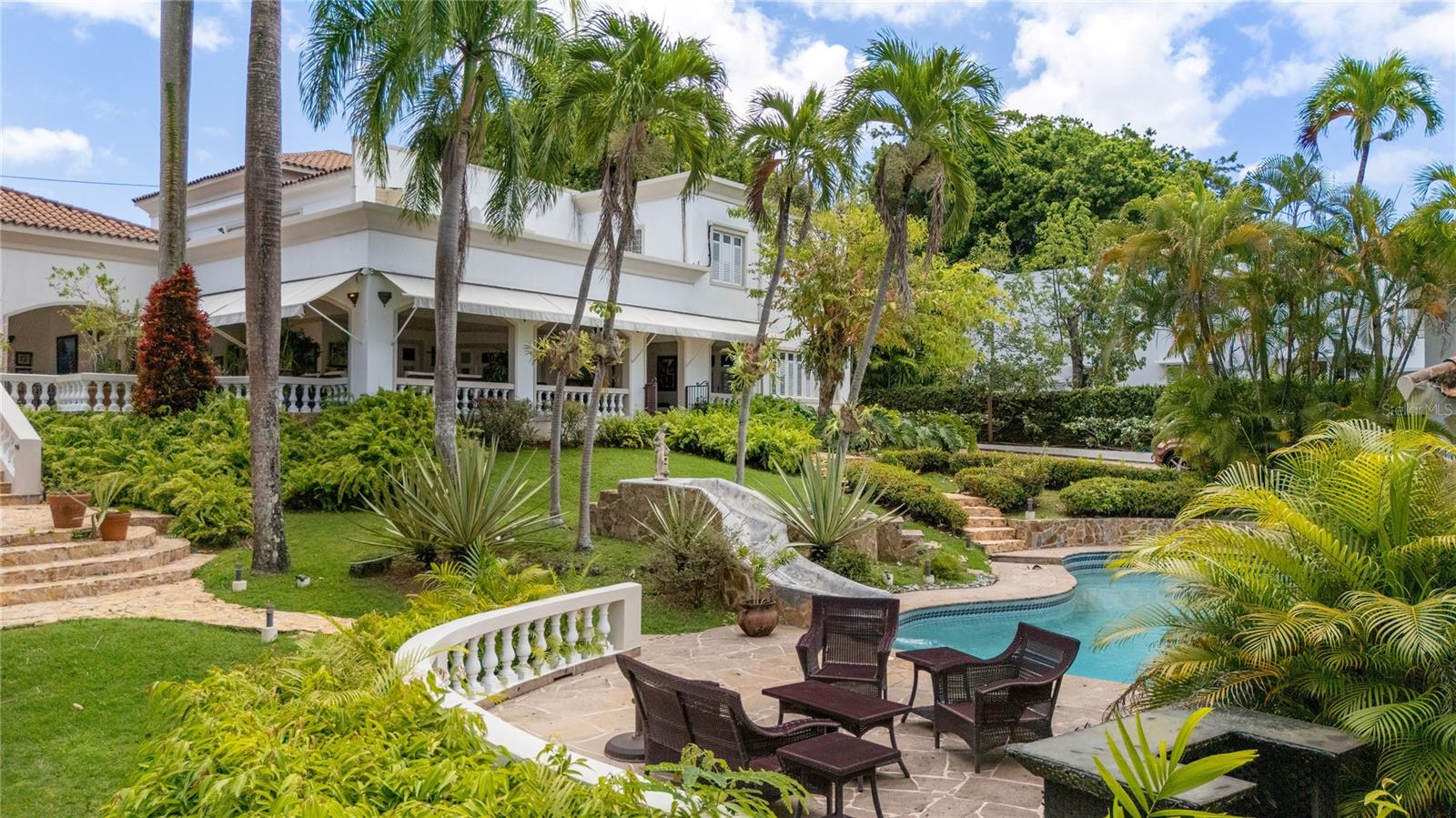 Welcome to one of Villa Caparra's most special homes, ideally located on prestigious A Street, the neighborhood's most desirable address. Set on an extraordinary 2,600-square-meter homesite surrounded by mature trees and lush tropical landscaping, this elegant Spanish Colonial estate offers a level of privacy and serenity that makes it easy to forget you're in the heart of Guaynabo.Originally built in 1960, the home underwent a thoughtful renovation and expansion in 2000 by a renowned Puerto Rican architect who beautifully preserved its original Spanish character while transforming it into a home designed for today's lifestyle. The result is a residence that feels timeless, welcoming, and effortlessly elegant.Offering approximately 10,180 square feet of total construction, including 6,359 square feet of interior living space, the estate is comprised of a 4,659-square-foot main residence and a 1,700-square-foot detached guest house and office.From the moment you walk through the front door, you're welcomed by soaring ceilings, beautiful red marble floors, graceful archways, and abundant natural light. The formal living and dining rooms open seamlessly onto expansive covered terraces, creating the kind of indoor-outdoor lifestyle Puerto Rico is known for. Whether you're hosting a celebration or simply enjoying a quiet afternoon surrounded by nature, every space feels warm, inviting, and connected to the outdoors.Upstairs, the main residence offers four spacious bedrooms and three full bathrooms, including a generous primary suite with oversized his-and-hers walk-in closets. At the heart of the home is a chef's kitchen with a large center island that naturally becomes the gathering place, complemented by a temperature-controlled wine cellar just steps away. The main residence also includes two interior powder rooms, a spacious laundry room, and an elevator serving both levels.The outdoor living spaces are equally inviting, featuring expansive terraces, an outdoor kitchen, two powder rooms, and a full bathroom serving the pool area. Just beyond the pool is a detached two-story guest house featuring a private office with its own full bathroom and storage on the first level, while the upper level offers an independent one-bedroom apartment with a private entrance and full bathroom—ideal for guests, extended family, or live-in staff.The home is equipped with a 40kW full-power generator, a 10,000-gallon underground water cistern, and a solar water heater, offering comfort and peace of mind year-round.Homes like this rarely come to market. Rich in character, surrounded by nature, and thoughtfully designed for entertaining, this is a property that offers a lifestyle unlike anything else in Guaynabo.