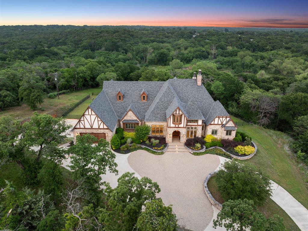Newly introduced at $4,749,000, this exceptional custom estate sits on 4.5 serene acres, backing to Corps of Engineers land for lasting privacy- offering rare seclusion in the heart of Flower Mound. A gated, tree-lined drive leads to a stunning villa inspired by the English countryside, thoughtfully designed for both everyday living and easy entertaining.
Built into the hillside, this unique home features a full level basement walk-out to the pool and spa, complete with two ensuite bedrooms, enormous entertaining space with game room, gym, wet bar, and dog grooming room. Upstairs, 20-foot beamed ceilings highlight the open-concept living area, anchored by a gourmet kitchen, dining space, and dual offices- all opening to a spacious covered veranda with a stone fireplace and outdoor kitchen, an ideal setting to relax and take in sweeping sunset views.
The primary suite offers a private terrace complete with a fireplace, offering direct access to the lower level outdoor backyard. Additional highlights include a large main-level ensuite guest room, a half court regulation basketball gym which can easily double as a pickleball court, Control4 home automation, oversized laundry and pantry spaces, foam insulation, Cat 5+ wiring, and a Class 4 roof. Room to expand with up to 1,600 additional sq ft. The 3-car garage currently serving as the basketball half court can easily be converted back, making for 5-car garages total. Sweeping circular drive provides ample extra parking.