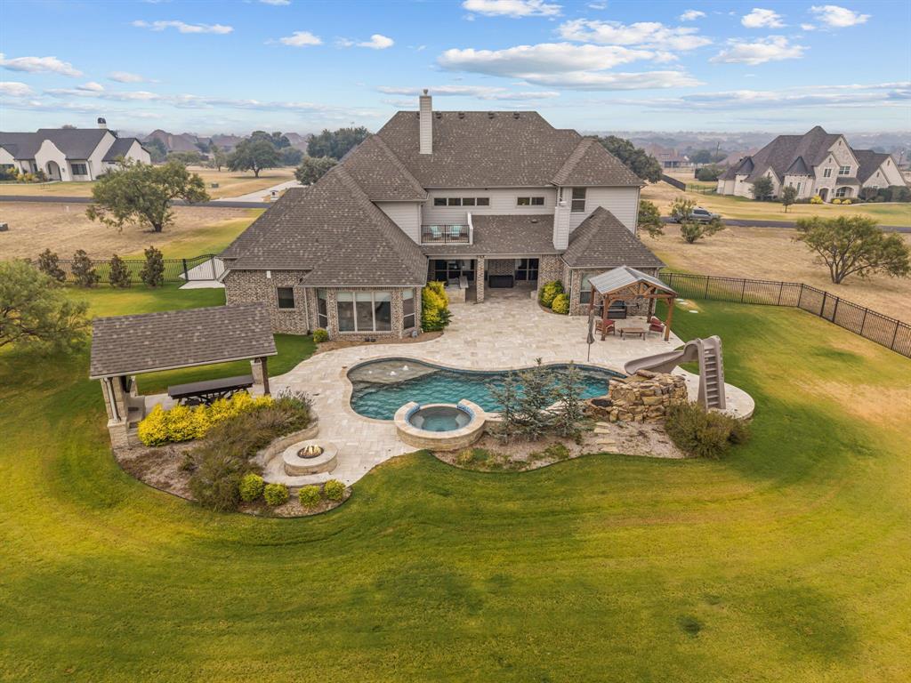 Set on 2.67 private acres in gated La Madera and Aledo ISD, this meticulously maintained 2019 Royal Crest Custom Home offers privacy, exceptional entertaining spaces and resort-style outdoor living. The backyard features a covered living area with fireplace and kitchen with built-in Blaze professional grill, plus a saltwater pool and spa with pebble finish, rock waterfall-grotto, slide, oversized tanning shelf, in-floor cleaning and Pentair smart controls. More than 2,500 sq ft of travertine patio, three covered patios, outdoor speakers, multiple gathering areas and three fire features create an exceptional setting for entertaining. Inside, soaring 23-ft ceilings and abundant natural light define the main living area. The chef’s kitchen features a 48-in KitchenAid Pro gas cooktop with griddle, double ovens and wine fridge. The first-floor primary suite includes automatic blinds. An expansive upstairs game room or secondary living space features a wet bar with fridge, freezer, kegerator, ice machine and sink, plus half bath and balcony overlooking the outdoor living areas. A tiered theater is wired for 9.2.4 Dolby Atmos. Additional features include 4 bedrooms, 3.1 baths, office, walk-in attic and insulated 4-car garage with epoxy floors. Systems include foam insulation, 3 HVAC units with 4 zones, R-8 ducts, recirculating hot water, well + water softener, dual hybrid water heaters, 250-gal buried propane tank, aerobic septic, smart sprinklers and generator wiring. Recent updates include a new Class 4 roof and well pressure tank. Built for safety, durability and connectivity, the home sits on a pier foundation anchored to bedrock and includes a hidden EF5-rated storm shelter panic room, wired security, 4K Lorex cameras and indoor-outdoor UniFi access points. Residents also enjoy La Madera’s private neighborhood lake and peaceful estate setting. No city taxes.