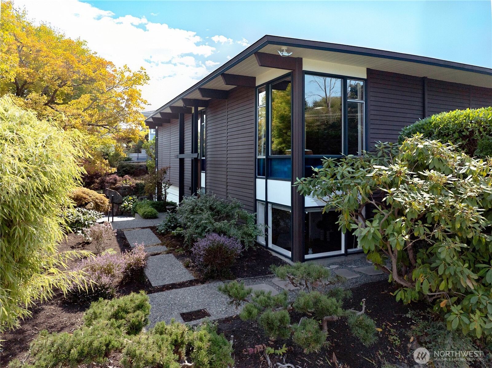 Once in a while, a truly special residence comes along. A place where time stands still, and every element exists in a harmony of design and purpose. A tribute to light and warmth, this custom architect-designed retreat embodies Mid-Century Modernism with an authenticity that is rare to find. Step into beautifully maintained interiors where the lines between indoors and out are blurred by luminous skylights, sweeping scenery, and full-length windows punctuated with colored glass. Slate and hardwood floors anchor elongated doors and open beam cedar-clad ceilings. Gather in the living area by a dramatic rock fireplace that invites you to relax and savor the sparkling waters of Puget Sound. Beyond, the open dining area and window-lined kitchen flow out to a private deck surrounded by mature Japanese maples. Make the most of sunny days in the generous yard with raised beds for your garden. Form meets function in this practical layout with all three bedrooms together on the main floor, plus an expansive daylight basement rec room and workshop/storage space that can adapt to your lifestyle. The oversized garage and ample driveway parking are every Seattleite’s dream. With Olympic Manor’s friendly serenity, festive holidays, and walkability to parks, schools, coffee, markets, and Golden Gardens, this is more than an address...it’s a lifestyle.