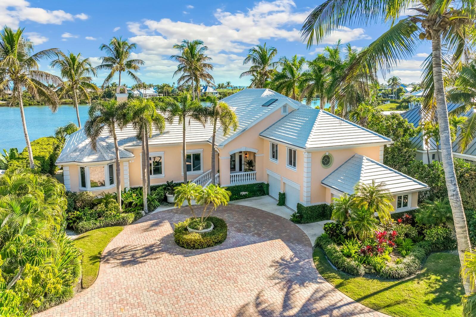 Bermuda-inspired coastal residence set within Boca Grande Isles, a private gated enclave overlooking sunrise views across Hole-in-the-Wall Bay. This spacious five-bedroom, four-and-a-half-bath home spans nearly 5,600 square feet of thoughtfully designed living space with classic architectural detail and relaxed island character.The exterior features clean stucco lines, white tile roof, and a gracious front porch. A circular paver drive leads to a maturely landscaped courtyard with palms and native coastal plantings. Inside, solid oak flooring, trowel-finished walls, and understated lighting create a calm backdrop for everyday living and entertaining. The living room includes a fireplace with custom carved woodwork and built-ins, and opens to a stone-floored sunroom framing views of the infinity pool and bay.The kitchen and keeping room form the center of the home, with granite counters, custom cabinetry, Sub-Zero refrigeration, Bosch dishwasher, double ovens, warming drawer, brick backsplash, and a wet bar with ice maker and wine refrigerator. The primary suite occupies the main level and offers dual walk-in closets, a sitting area, and a well-appointed bath with soaking tub and walk-in shower. Four additional bedrooms include two private guest suites, and an elevator provides convenient access to all levels.Outdoor living unfolds over terraced spaces surrounding the infinity pool, set along the bay and sheltered by mature landscaping. An oversized garage accommodates two vehicles plus a golf cart, with storage for kayaks, bikes, and beach gear.Located minutes from Boca Grande’s historic village and Gulf beaches, this property combines privacy, scale, and waterfront living in one of the island’s most desirable settings.