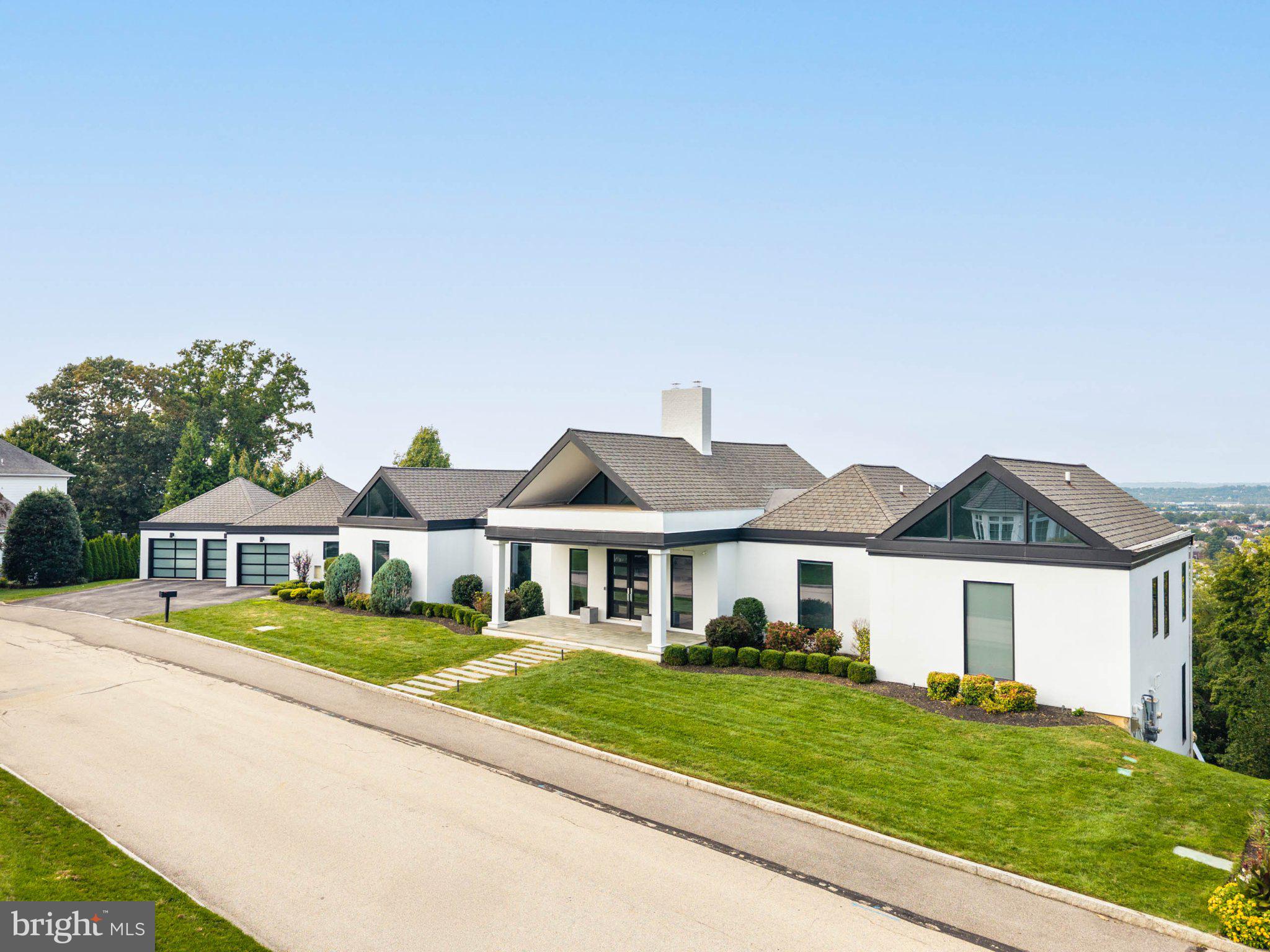 Set above the landscape in Gladwyne, this dramatic contemporary residence is defined by extraordinary, miles-long views and an interior that feels equally expansive. Minutes from the Main Line’s conveniences yet seemingly worlds apart, the home pairs a crisp, sophisticated palette with exceptional scale, thoughtful design and an almost constant connection to the horizon.Double glass doors open to a soaring two-story foyer, where wide gallery-like halls and European white oak floors extend east and west. A dramatic two-sided gas fireplace, finished in painted brick and rising a story and a half, acts as a sculptural divide between the entry and great room, offering fire and sightlines to both spaces. Beyond, the great room opens through broad sliding glass doors to a deep terrace, effectively extending the living space outdoors. From here, distant rolling hills give way to city lights, creating a backdrop that changes beautifully from morning through evening.Beyond the great room, the dining room continues the home’s nuanced palette of whites and warm natural materials. Far from a space reserved only for occasion, it is designed to be used and enjoyed. As daylight fades, the room becomes particularly compelling, with illuminated views beyond and doors opening directly to the shared terrace for cocktails, dinner or evenings outdoors.The kitchen is striking in its restraint. Gleaming lacquered white cabinetry is paired with substantial quartzite surfaces, gently veined for subtle movement and warmth. An oversized island provides generous room for seating, preparation and display, while a professional suite of Thermador appliances includes full-size side-by-side stainless steel refrigerator and freezer columns. Against the polished cabinetry, a natural wood barn door concealing the pantry introduces an organic counterpoint. An informal dining area sits just beyond, as does the family room, open enough to feel connected to the kitchen yet sufficiently removed to allow several conversations, and several generations, to comfortably coexist.Privately positioned to the right of the foyer, the expansive primary suite continues the home’s high ceilings, European white oak floors and serene aesthetic. Two exceptional dressing rooms provide extensive hanging, drawer and folded storage; one is large enough to incorporate a full desk beneath a bank of windows, creating a beautifully integrated private workspace.Large-format tile introduces the primary bath, where two wall-hung vanities with stone tops, a soaking tub beneath the windows, private water closet and generous steam shower with body sprays and bench create a space that feels both architectural and indulgent. Southern views remain present throughout.Equally considered are the practical spaces: two powder rooms, a substantial mudroom, direct access to the three-car garage and even an elevated dog wash. Nothing feels incidental. Each element has been thoughtfully incorporated into the architecture of the house.Two separate staircases descend to an expansive garden level, where the sloping site becomes one of the home’s greatest assets. Rather than reading as a traditional lower level, these rooms open directly to another deep terrace and the landscape beyond. A central fireplace anchors a generous family room with multiple gathering areas and ample space for billiards, ping-pong or both. A built-in bar with glassware storage and refrigeration allows entertaining to move effortlessly between floors.Four additional bedrooms are found here, two with en-suite baths and two sharing a central bath while retaining individual vanities. A dedicated gym completes the level. Bright, expansive and finished with the same attention given to the main floor.The result is a home of unusual confidence: contemporary without feeling austere, glamorous without excess and exceptionally well suited both to large-scale entertaining and everyday life.