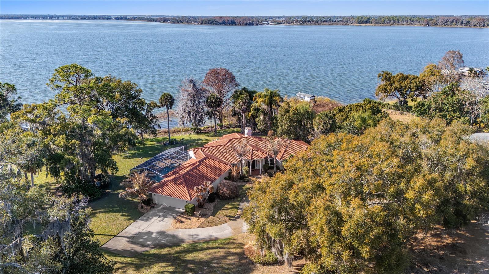 Envision the arrival of your private lakefront estate on the storied shores of Lake Dora, the crown jewel of the Harris Chain of Lakes, a vast and celebrated waterway offering endless miles of lock and river cruising for days defined by leisure and adventure. Life on the lake unfolds at a gentler, more inspired pace as mornings begin with coffee on the dock while mist rises from the water, afternoons invite boating, paddleboarding and fishing along scenic shorelines, and evenings conclude with unforgettable sunset cruises beneath painted skies. Just moments away, the charm of Mount Dora enriches the lifestyle even further. Known for its storybook streets, vibrant arts scene and year-round calendar of festivals, residents enjoy boutique shopping, waterfront dining, antique markets, boat shows, food and wine celebrations, and lively community gatherings that make every season feel like an occasion. Across a remarkable eight-acre parcel, 6136 Dora Drive commands hundreds of feet of pristine shoreline, ideally oriented to the west for breathtaking sunset vistas that paint the water in gold each evening. This extraordinary canvas invites limitless possibilities such as design a grand equestrian compound, cultivate a private orchard, create an inspired guest residence with an expansive collector’s garage or explore opportunity to thoughtfully subdivide. Within a canopy of mature trees, a sprawling single-story residence spans approximately 4,000 square feet, offering three bedrooms, two and a half baths, walls of windows, a screened pool and spa, and a newer tile roof. Completing this rare offering is an enclosed boathouse with private doors, an increasingly scarce luxury along the chain. With no HOA restrictions and boundless space to create, this is a legacy lakefront setting where vision meets lifestyle, a place where lake days, sunset evenings and timeless charm of Mount Dora define life at its most exceptional.