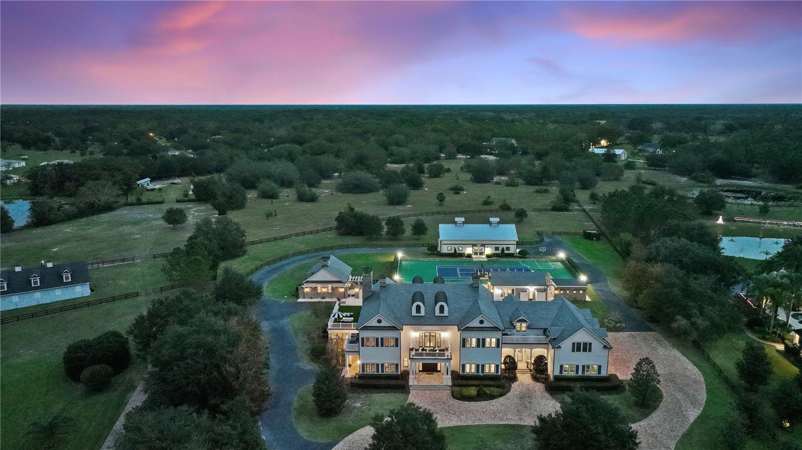 This home isn’t for the faint of heart … it’s a showstopper.  Just under 14,000 ft.², 5 buildings, 13.75 neat & tidy acres in the double gated enclave of Mills Cove. 30 mins to DT Orlando, less than an hour to the beach, 10 minute helicopter ride to the airport, yet you’ll feel like you’re on your country estate.The quintessential entertainment home radiates quality with a hyper-luxe moment around every bend. The undeniable quality of this developer’s personal dream home, is evident from the moment you cross the threshold into the 2014 Parade of Homes Custom Showcase GRAND AWARD WINNER by Hill Gray Seven’s Gregg Hill.  Set back an acre, guests are greeted in neo-colonial style, with the pomp & circumstance one expects for heads of state. Travel around from either gate, to the front stone entry or round either corner to the property edges, then head back to the two bedroom guest cottage w/ stable beyond, or (on the other side) past the sports complex/tennis/pickleball/basketball courts to the stables. Saunter in from the grande front portico and you’ll be transported to an era when hospitality, ease & decorum were to be expected.  Once inside, you’ll be warmly welcomed by a 2 story entrance gallery flanked by (4) 13’ double arched custom mirrored niches. Left to the 2 story, wood paneled study & fireplace w/separate entry, a British inspired sports pub/trophy room, a powder room w/hand laid marble mosaic tiles. Head right you’ll find the Fibonacci-esque spiraling staircase anchored by the game playing studio. Entertainment meets unencumbered formality.  The grande living room, fireplace & hearth is opposite the kitchen w/ triple thick rockface edged slab, 12’x 16’ kitchen continent. Sweeping views of the resort sized aquatic playground just beyond.  Kitchen adjacent are breakfast & wine tasting rooms, showcase pantry, craft room, 2nd powder room, mud room, doggy bathroom (w/shower, separate tub, washer/dryer & pups potty access). The sunroom hosts formal dining & music conservatory anchored by it’s weighty brick fireplace & hearth.  Upstairs, the primary bedroom, faux turf deck for lounging, improving your putting game, or surveilling your dominion.  The primary bath is a work of art with hand laid leaf marble mosaic tile harkening to the 11th century Moorish Maallem’ craftsmen.3 en-suite bedrooms, a bunkhouse, billiards room, home theatre, 2 additional turfed decks, a 3rd story bonus room provide space for everyone to “do their thing”.  A 2nd staircase, provides additional access & flow.  The pool accommodates your entire guest list, sunken summer kitchen & swim up bar serves meals under sunny skies, or sur la nuit étoilée. A 2 bdrm guest cottage w/ kitchen & fireplace keeps guests comfy & well accommodated.  Fitness studio overlooks pool & tennis courts, tennis viewing platform easily seats 12.  The freestanding yoga/dance studio adjoins the tennis courts and al fresco lounging patio is an ideal vantage point to watch a good match. The payoff for good workout is a relaxing aromatherapy steam shower. The 5 stall aisle barn & tack room is immaculate, grooming stations, trainer’s studio apartment, full length hayloft/storage. Paddock for turning out or a manège, cleared acreage behind the stables is for riding, establishing an orchard, or both.  Familiar and comfortable, even the deer feel relaxed here, meandering without hesitation.  So will you. Broker is Lic'd Real Estate Agt. MLS details to be verified by buyer