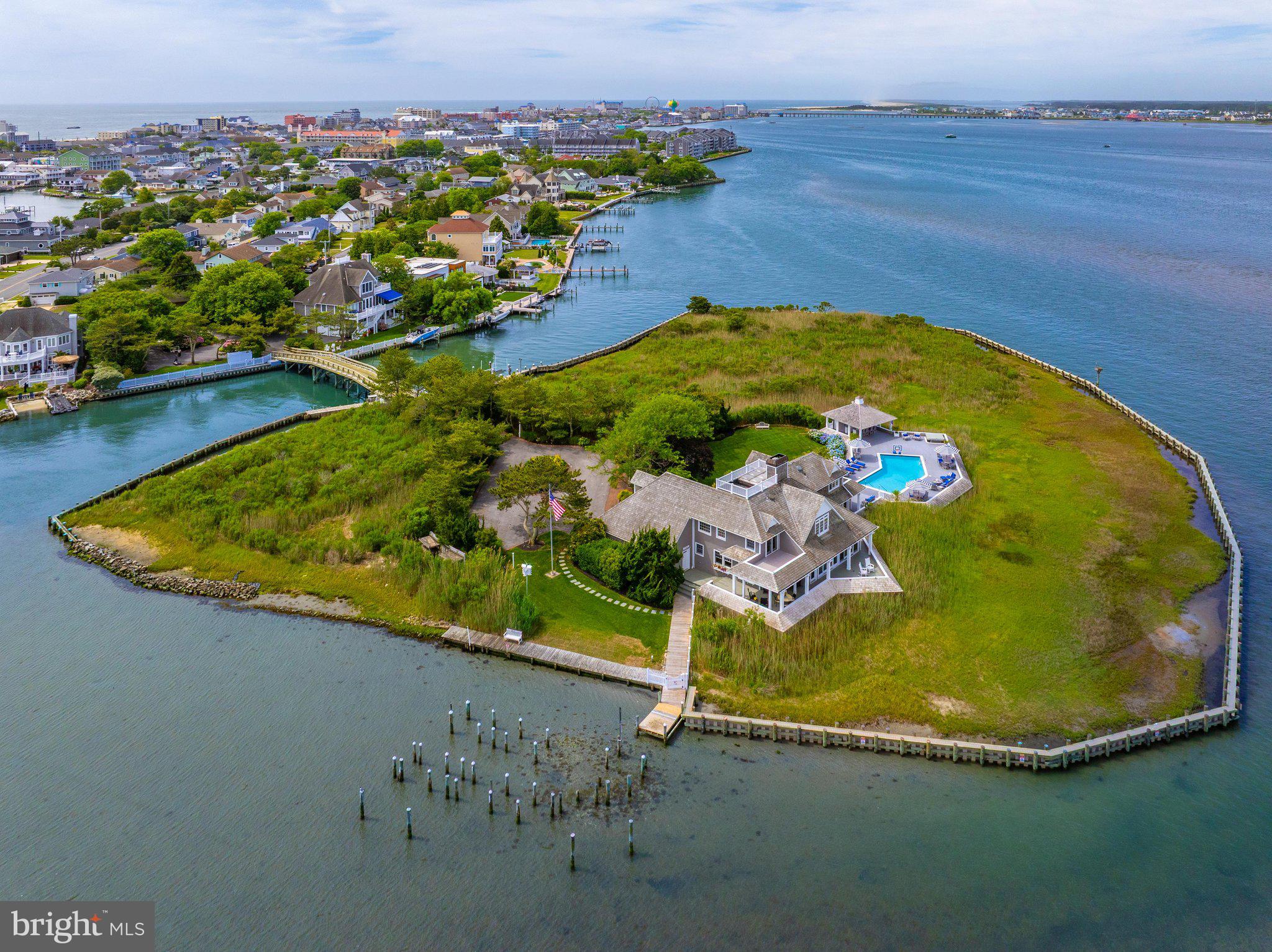 Private Island at 1546 Teal Drive, Ocean City, Maryland is a truly unique offering — the bay's only island estate. Included is an adjacent buildable waterfront lot just inside the entrance gates before crossing the private bridge. The improved .29-acre parcel features approximately 95 feet of bulkheaded shoreline with boardwalk, paved driveway, irrigation, and landscaping, offering flexibility for a future guest house or single-family residence, additional parking, or expanded waterfront enjoyment. Located off 15th Street in downtown Ocean City, the property offers seclusion and accessibility, 3 blocks to the Boardwalk and beach, minutes to the Ocean City Inlet and Assateague Island by boat, and a block from Harbor Island, home of the world-renowned White Marlin Open. Hidden behind a brick wall and accessed through a 6-foot gate, the arrival continues across a private 120-foot vehicular bridge from Mallard Island. The island encompasses more than 3 acres of native marshlands and coastal landscape with panoramic views of the open bay, the Rt. 50 Bridge to the south, Rt. 90 Bridge to the north, and the rare combination of breathtaking sunrises and sunsets across the water. Architecturally, the home is an iconic shingle-style residence inspired by Nantucket architecture, with exterior renovations and enhancements by renowned Lyman Perry Architects Ltd. of Nantucket, Massachusetts. The approximately 5,000-square-foot home features custom woodworking, Carlisle wide-plank hand-finished pine flooring, new Andersen doors throughout, approximately 3,250 square feet of wraparound decking, and expansive water views. Upon entering, a dramatic 24-foot atrium features a 12-foot by 15-foot overhead skylight and floor-to-ceiling Coral Rock fireplace feature connecting the surrounding living spaces. Designed to maximize open bay views throughout, the main living area features custom built-in cabinetry and flows into the separate dining and gathering spaces. The kitchen/family room creates an inviting area for entertaining, with custom solid birch painted cabinetry by Premier Cabinetry of New Holland, Pennsylvania, and a 5-foot by 8-foot teak island countertop. A custom wood spiral staircase provides secondary access between the main and upper levels. The main level also includes a home office with built-in cabinetry and pull-out file storage, plus an additional guest bedroom and full bath. Upstairs, an open bridge hallway overlooks the atrium while separating the primary suite from two guest bedroom suites. The four-room primary suite includes a bedroom retreat with bayfront deck, sitting/entertainment/dressing room with vaulted ceilings, custom cherry wood built-ins and four closets, en-suite bath with double vanity and dressing table, and a second dressing room with vaulted ceilings, four double-door deep closets, and an arched window with window seat dividing two built-in tall chests. Two additional bedrooms each feature en-suite baths and private decks. Outdoors, an island retreat features a pool and spa by Sea Glass Pools, expansive decking, gazebo bar with cabinetry and quartz countertops, full bath with shower, docking, 10,000-pound boat lift, and fish cleaning station. Significant recent investments include bulkhead improvements, reinforced helical pilings, bridge renovations, active security and surveillance systems, and maintained structural and mechanical systems. Surrounded by abundant birdlife and endless open-water vistas, this distinguished estate embodies timeless elegance.