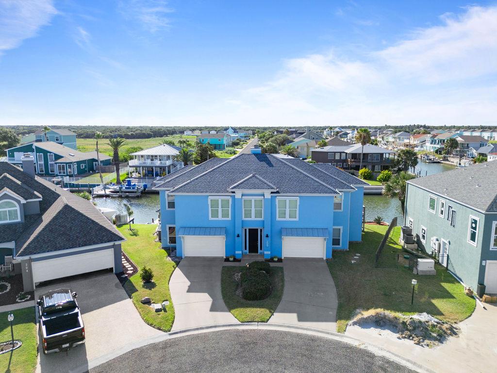 Aerial view of a blue waterfront home on Reserve Lane in Rockport, Texas, showcasing nearby houses, palm trees, and a boat dock, emphasizing the coastal living experience.