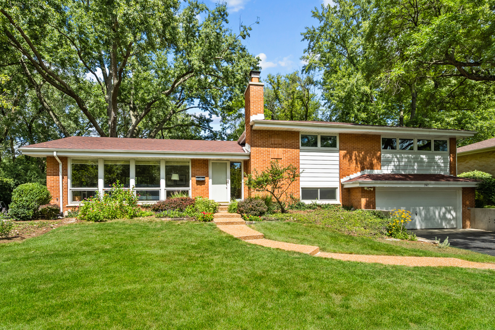 Tucked away on a private, tree-lined lot, this beautifully updated four-bedroom, two-and-a-half bath home blends mid-century character with modern comfort. Expansive windows frame serene views of nature and fill the interior with natural light, while beautiful hardwood floors and thoughtful updates create a fresh and inviting atmosphere throughout. The main level opens with a spacious living room anchored by a wall of windows, flowing seamlessly into the dining room and out to the patio for easy entertaining. A bright kitchen features quartz countertops, crisp cabinetry, stainless steel appliances, and an adjoining breakfast nook with peaceful views of the backyard. Just steps away, a large family room with a fireplace offers a comfortable retreat with sliding doors to a lower patio. A powder room and large laundry/mudroom lead to the attached 2-car garage. Upstairs, you'll find a generous primary bedroom with an ensuite bath, three additional bedrooms, and an updated hall bath with dual sinks. The finished lower level extends the living space and is perfect for a gym, workshop, or recreation room. Outside, the home is surrounded by mature trees and lush landscaping, offering a rare sense of privacy. Multiple patios overlook the deep yard, perfect for gatherings, gardening, or quiet afternoons. Set on a quiet street yet close to schools, parks, town, and the lakefront, this is the perfect place to call home.