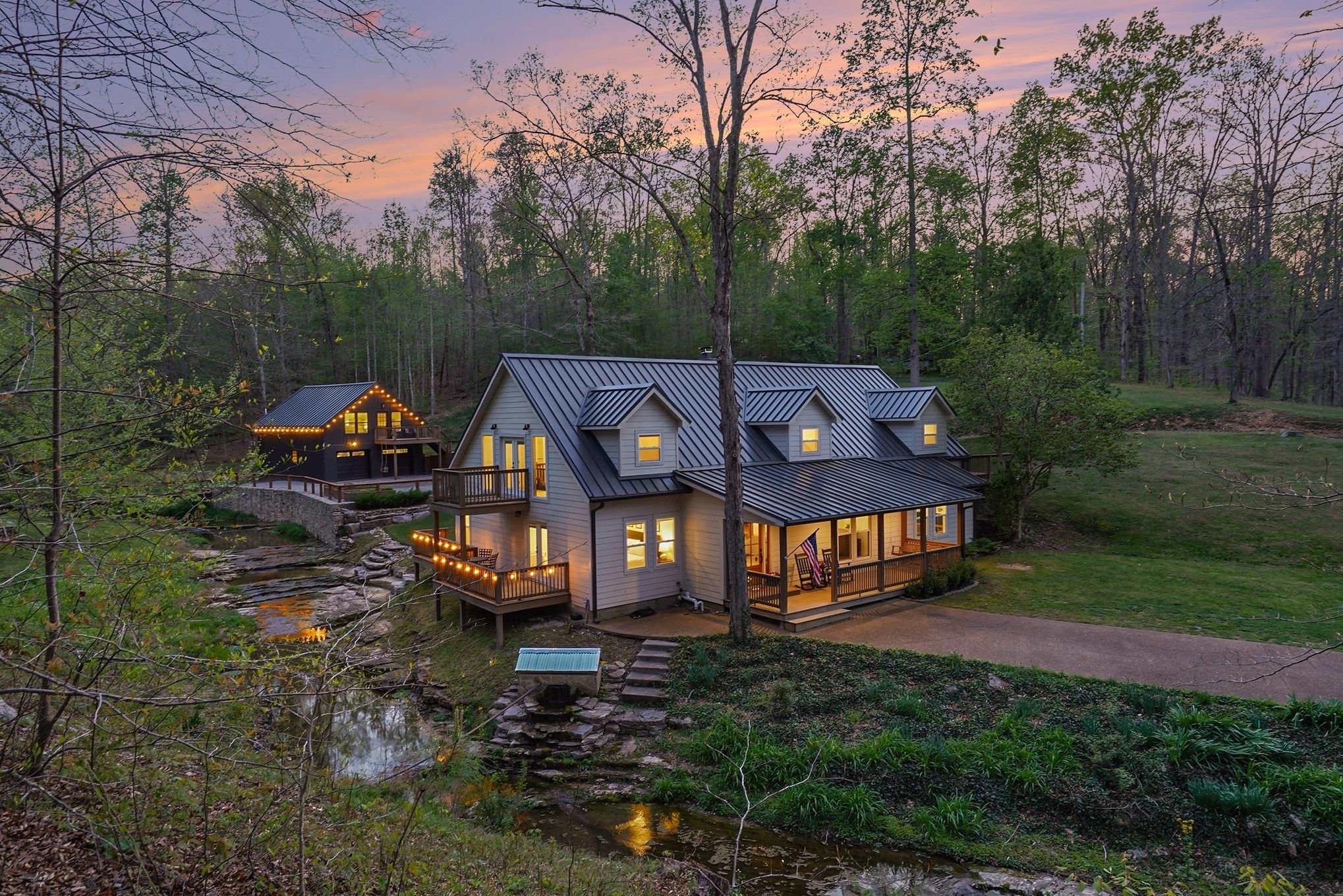 Tucked away on 4.59 breathtaking creek-front acres in Williamson County, this one of a kind property offers an exceptional blend of privacy, natural beauty and versatility. From the moment you turn onto the tree-lined drive, the setting feels special, with the driveway meandering alongside Caney Fork Creek as it leads to the home. The sound of cascading water creates a peaceful backdrop throughout the property and can be enjoyed from three decks, a welcoming front porch and private backyard. The home is filled with warmth and character, featuring beautifully curated finishes, custom built-in dining seating and abundant windows framing picturesque views. Adding tremendous flexibility is the detached 2-car garage with a fully finished 1-bedroom apartment above, currently operating as an income-producing Airbnb that is ideal for guests, extended family, a private office or continued rental use.  The 4.59 acres offer a beautiful balance of open space and mature woods with room for gardening, recreation and private walking trails. A charming bridge crosses the creek to a fire pit area tucked among the trees, creating a storybook setting for gatherings and evenings under the stars.Set well off a quiet, low-traffic country road, the property feels wonderfully private while remaining just minutes from everyday conveniences and an easy drive to Leiper’s Fork and downtown Franklin and Nashville. Recent improvements include a new roof (2024), backyard fencing, whole-home water filtration system, enclosed storm shelter with new steel door, custom dining built-ins and a new electrical panel in the workshop. A truly special Williamson County retreat!