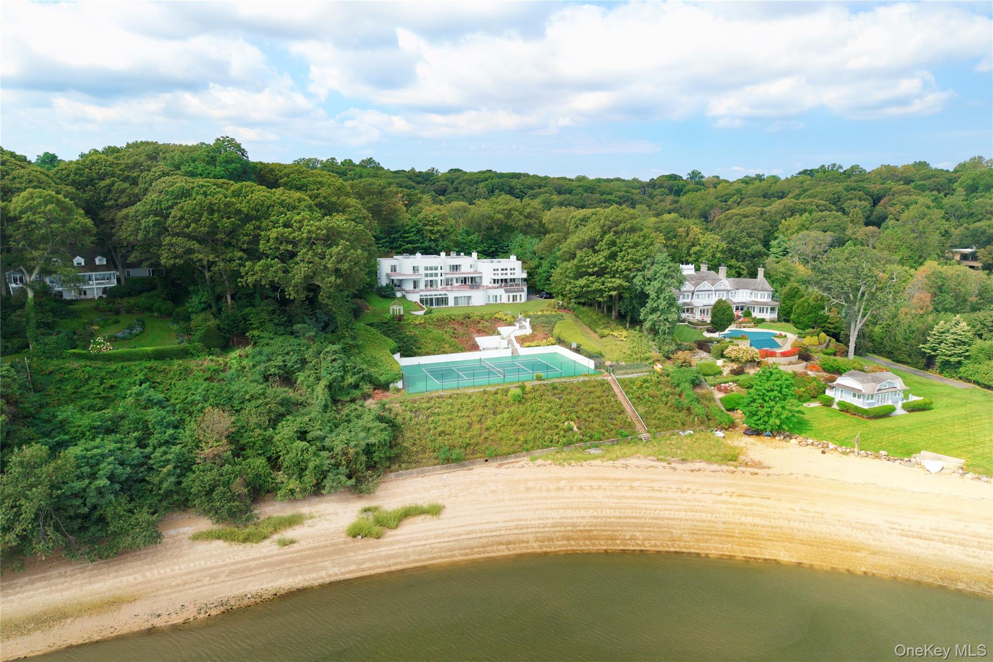 Experience the ultimate in waterfront living with this extraordinary 9,000+ sq. ft. estate set on two sweeping acres with 200 feet of private shoreline in Oyster Bay. Designed for both grand entertaining and everyday comfort, the residence offers 5 bedrooms, 6.5 baths, and 4 fireplaces, all supported by modern essentials including a full-house generator and security system.Step inside the dramatic entry foyer where light and space unfold into expansive living areas with floor-to-ceiling windows framing panoramic water views. The main level is highlighted by a sophisticated living room, formal dining room, and a versatile lounge/meeting space. A library with its own bath provides a perfect retreat or guest option. The chef’s kitchen impresses with granite counters, custom cabinetry, professional appliances including three ovens and two dishwashers, and an adjoining laundry/mudroom. A spectacular 1,600 sq. ft. great room with a wet bar and wood-burning fireplace anchors the first floor.Upstairs, three ensuite bedrooms each open to private balconies overlooking the water. The primary suite is a true sanctuary featuring a fireplace, spa bath with dual vanities, and an oversized walk-in dressing closet. Outdoor living is elevated with more than 1,800 sq. ft. of wraparound decking, a tennis court, gazebo, and direct water access for kayaking, jet skiing, and more.The lower level extends the lifestyle with an indoor pool and hot tub, an entertainment space with wet bar, a full guest suite with private entrance, abundant storage, and a 4+ car garage. With meticulous craftsmanship and unparalleled amenities inside and out, this residence is not just a home—it’s a private waterfront resort.
