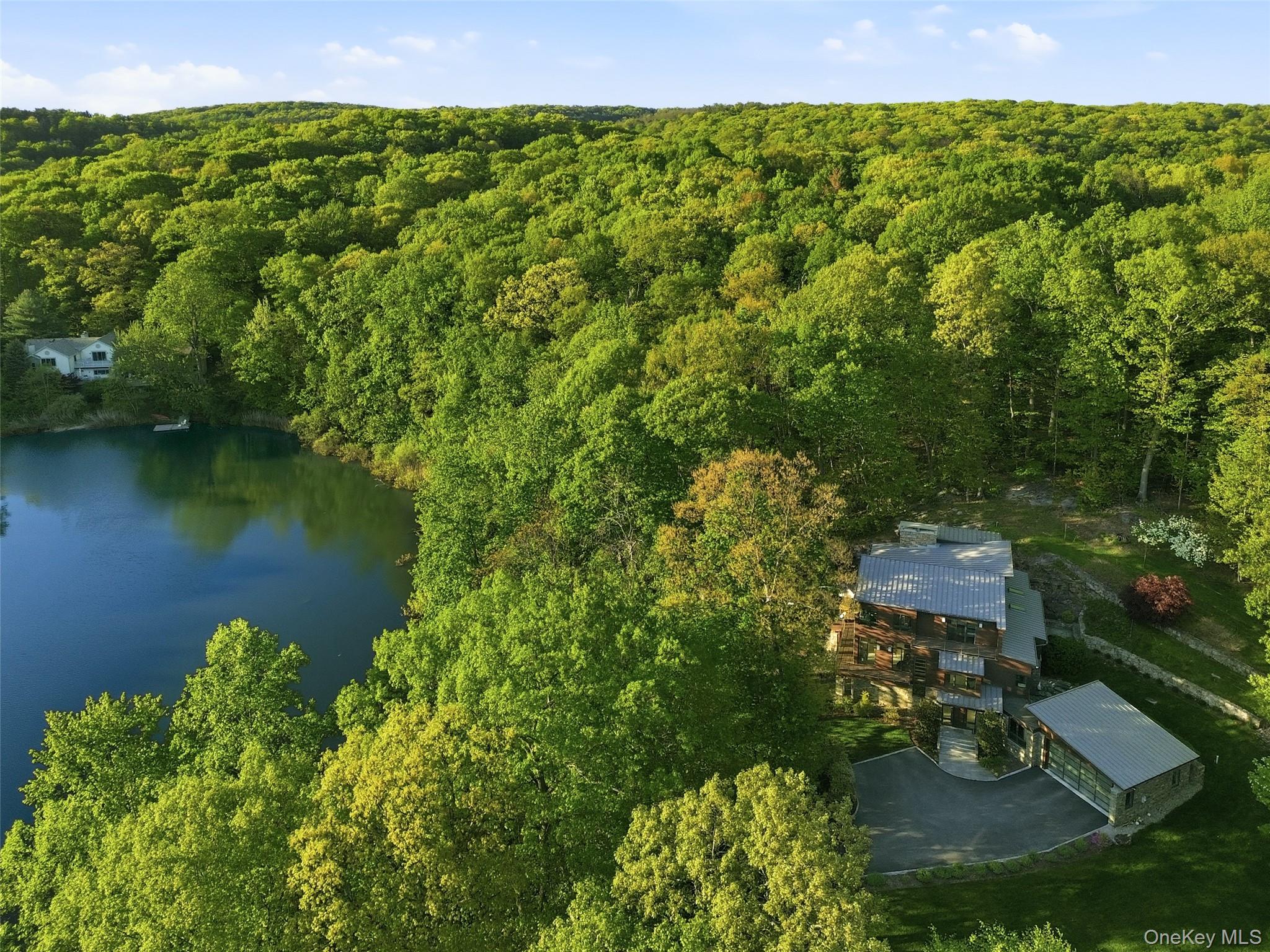 Modern Luxury Meets Lakeside Serenity. Experience a rare blend of privacy and year-round recreation in this custom-built Armonk home set directly on the water’s edge. Designed for effortless living, it offers kayaking, swimming, and peaceful mornings from your private dock. Outdoors, a lakeside fire pit, outdoor shower, and secluded hot tub are framed by panoramic water views. The home is thoughtfully designed to blur the line between indoors and out. Floor-to-ceiling windows and bi-fold glass doors open to expansive decking and striking dry-stack stonework, creating seamless flow. Inside, wide-plank flooring and a dramatic two-story great room with a floor-to-ceiling stone fireplace establish a warm, refined atmosphere. The space connects to a chef’s kitchen with premium appliances, walk-in pantry, and wine refrigerator. The primary suite is a private retreat with its own fireplace, deck access, and spa-like bath. Upstairs, three bedrooms surround a flexible lounge ideal for a media room or play space. The finished entry level includes a home gym, theater, and large mudroom. Minutes from town yet completely private, this exceptional property offers a true modern lakefront lifestyle.