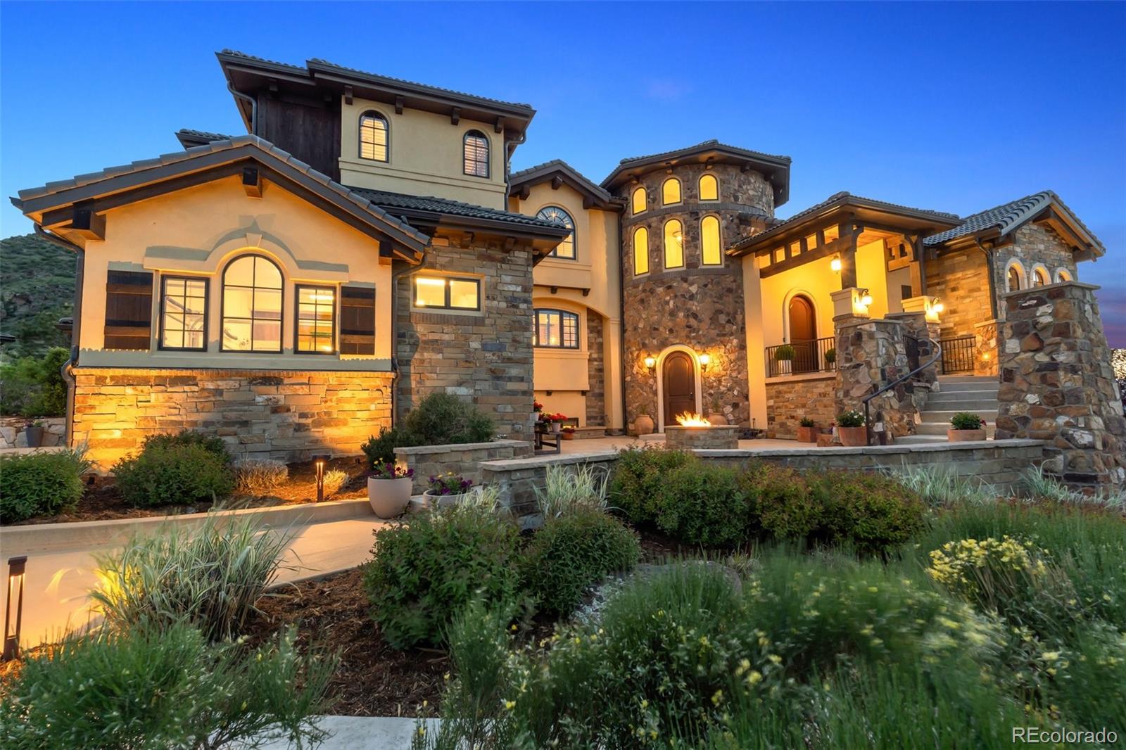 Welcome to Ravenna – Denver’s Premier Gated Golf Community. This Italian-inspired, three-level custom home is nestled on one of Ravenna’s few forested lots, offering exceptional privacy, timeless elegance, and panoramic views of downtown Denver and Chatfield Reservoir. A tree-lined driveway leads to the grand entrance, where a stone-vaulted foyer opens into expansive living, dining, and kitchen areas beneath a soaring 25-foot beamed ceiling. A commanding stone fireplace anchors the space, complemented by dark oak hardwood floors and crowned with Restoration Hardware chandeliers. A wall of southwest-facing windows frames unobstructed views from the backyard’s wooded gully, across the 3rd green, and into the rolling hills of Pike National Forest. The main-floor primary suite shares in this dramatic vista, with direct patio access, built-in bookshelves, an electric fireplace, and a luxurious spa-like bath featuring a soaking tub and steam shower. Adjacent is a stately study with fireplace and custom built-ins, completing the private main-floor wing. Upstairs, two guest suites and a spacious family/playroom provide ample accommodations, while the walk-out lower level is designed for multigenerational living—featuring three ensuite bedrooms, a home theater, generous living/play space, and a private patio with downtown Denver views. Additional highlights include smart home controls (garage, HVAC, security, doorbell), a gas firepit, Pella windows, Hunter Douglas electric shades, custom drapes and shutters, and an oversized four-car garage with ceiling racks, 7-ft cabinets, and a 4-passenger golf cart with new lithium batteries. This one-of-a-kind property is protected by a 400-ft bespoke stone wall and offers resort-style living with access to hiking, boating, golf, and fine dining—right at your doorstep.