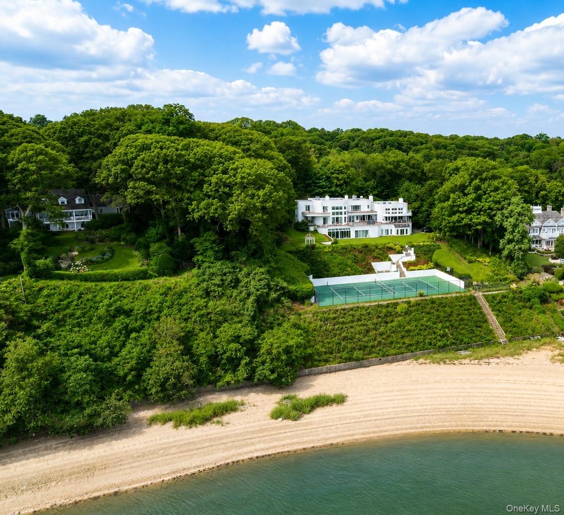 On the tranquil shores of Cove Neck in Oyster Bay, this extraordinary waterfront estate offers something increasingly rare: two private acres, 200 feet of shoreline, and panoramic views of the Long Island Sound that greet you from virtually every room in the house.Set just minutes from the charming villages of Oyster Bay and Huntington, with their acclaimed restaurants, boutique shopping, and vibrant waterfront dining, and fifteen minutes to the LIE, this is a property that offers the serenity of a private resort with the convenience of everything the North Shore has to offer. Hamptons Living Closer to NYC.The residence spans more than 9,000 square feet and was designed for those who want to live at full scale. A dramatic entry foyer opens into expansive living areas anchored by floor-to-ceiling windows framing the water. The formal living and dining rooms, a sophisticated lounge, and a 1,600 square foot great room with wet bar and wood-burning fireplace create a seamless flow for entertaining. A library with its own bath offers a quiet retreat on the main level.Upstairs, three ensuite bedrooms each open to private balconies overlooking the Sound. The primary suite is a sanctuary of its own, with a fireplace, spa bath with dual vanities, and an oversized walk-in dressing closet. Every room. Every view.The chef's kitchen is equipped with granite counters, custom cabinetry, and professional appliances including three ovens and two dishwashers. The lower level extends the lifestyle further with an indoor pool and hot tub, an entertainment space with wet bar, a full guest suite with private entrance, and a 4-car garage.Outside, more than 1,800 square feet of wraparound decking, a tennis court, a gazebo, and direct water access for kayaking and jet skiing complete a property that operates entirely on its own terms.This is waterfront living on the North Shore at its most complete.