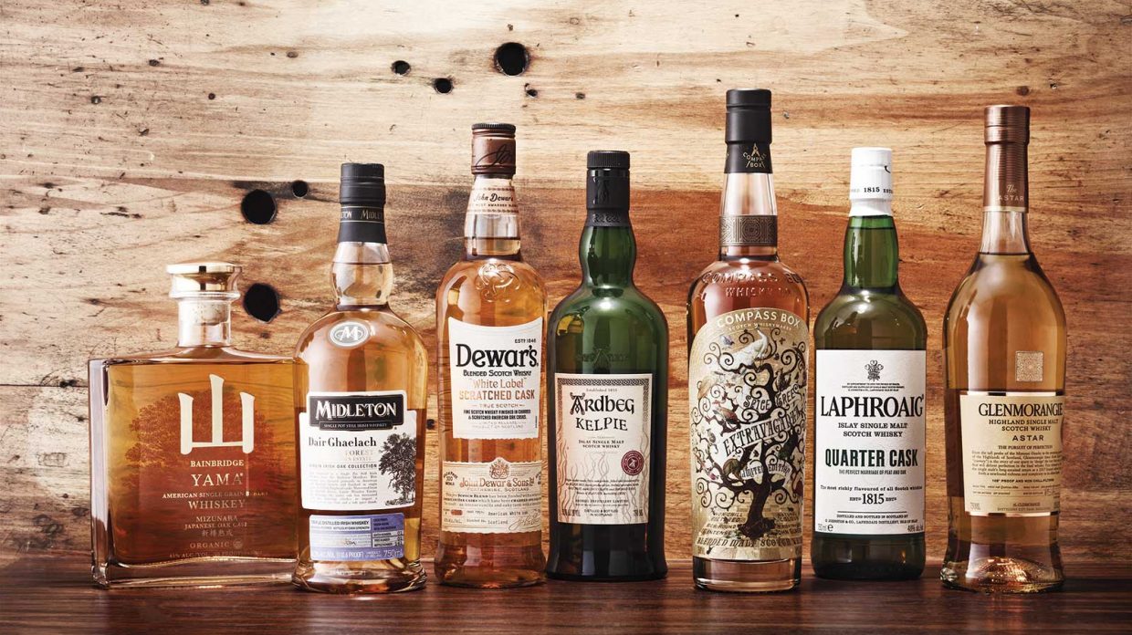 Several bottles of whiskies matured in custom-made casks rest on a wooden surface in front of a wooden wall.