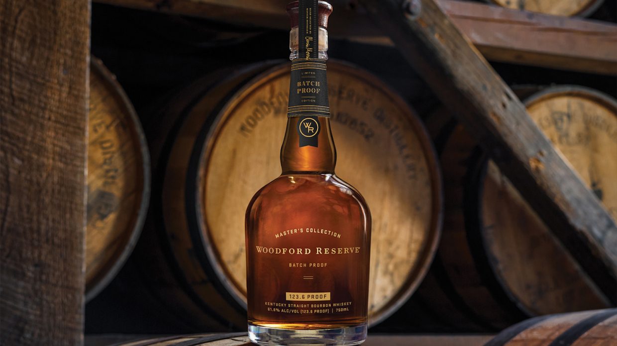 A bottle of Woodford Reserve Batch Proof (2020 Edition) sits on top of a whisky barrel, with several barrels visible behind it.