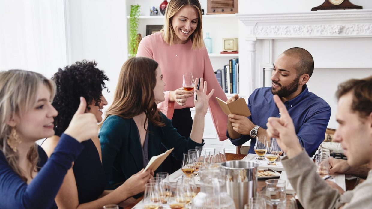 A group of six friends sits around a table to enjoy a whisky tasting.