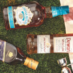 Bottles of blended scotch—Johnnie Walker Blue Label, Dewar's White Label, and Chivas Regal Mizunara—rest on their sides, pictured from a bird's-eye view, atop what appears to be close-cropped grass, next to a red and white plaid-patterned picnic blanket, along with a wooden-handled metal jigger, and two rocks glasses containing ice and what appears to be whisky cocktails; the brim of what appears to be a straw hat also is partially in view to the lower left corner of the photo.